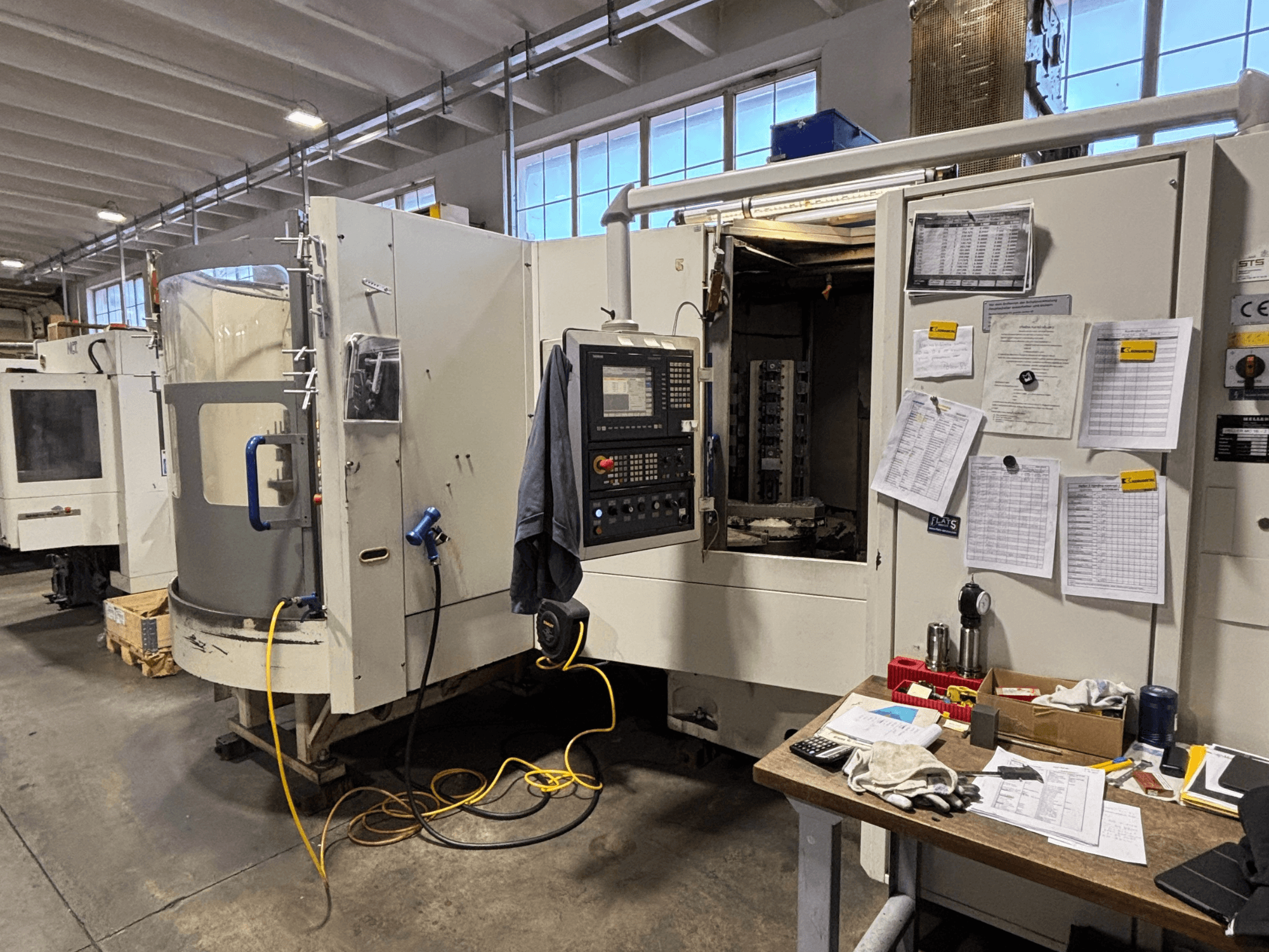Верстат з ЧПУ в майстерні, вид спереду, з панеллю керування та документацією збоку; інструменти та матеріали на столі.