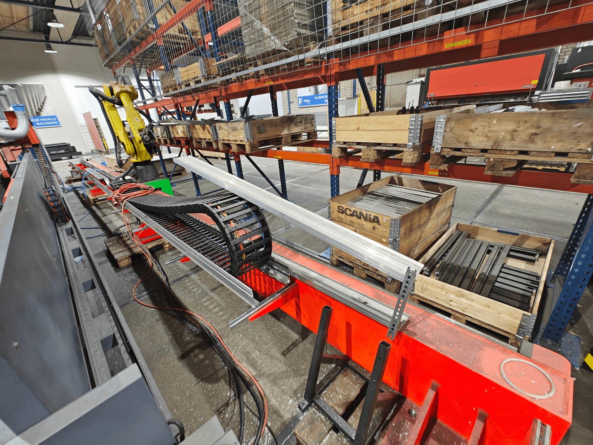 Роботизована рука на робочому місці, видно помаранчеві та чорні кабелі, з ящиками Scania та алюмінієвими профілями у заводських умовах.