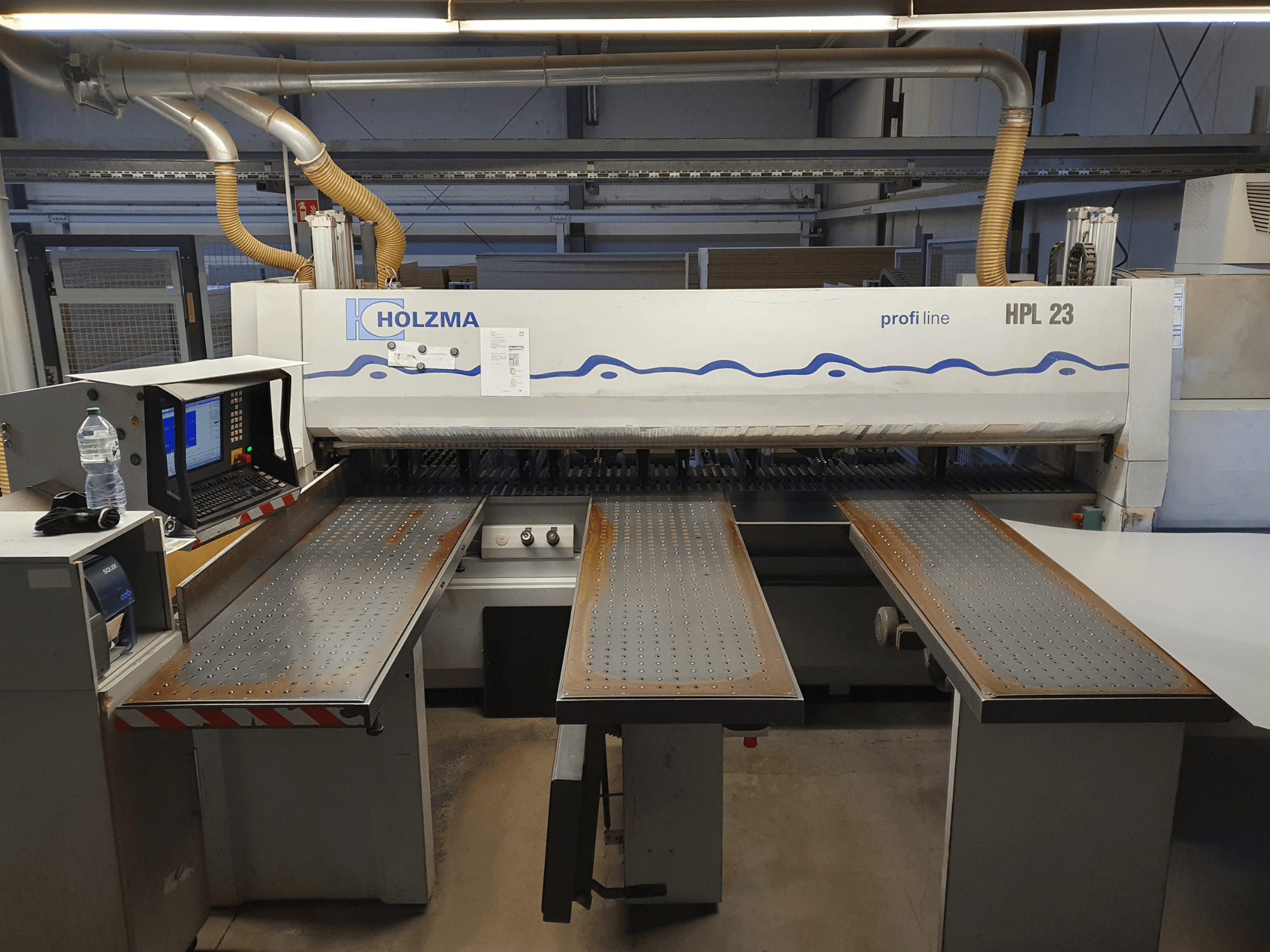 Розкрійний верстат HOLZMA HPL 23 показаний спереду, з трьома перфорованими столами, комп'ютерним терміналом і пляшкою води поруч.