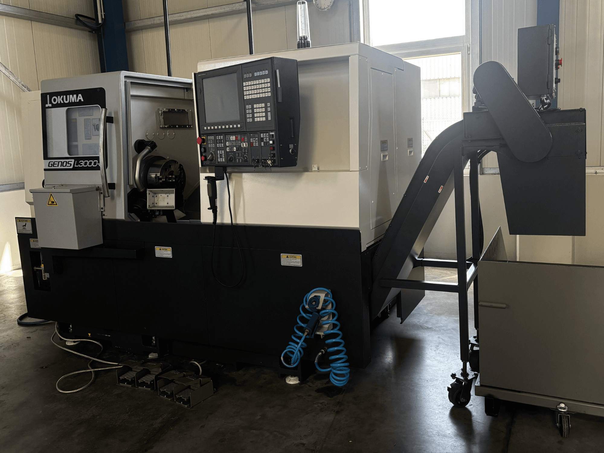 Горизонтальний токарний верстат Okuma GENOS L3000-e, вид спереду, на якому видно панель управління, конвеєр та зону шпинделя.