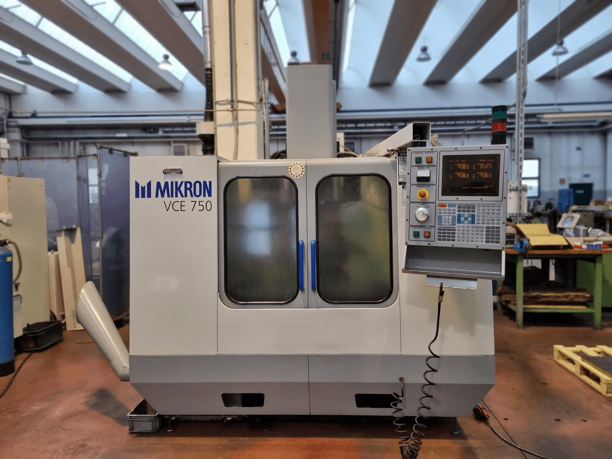 Білий верстат з ЧПУ Mikron VCE 750 в майстерні, вид збоку, з цифровою панеллю керування та великими скляними дверима.