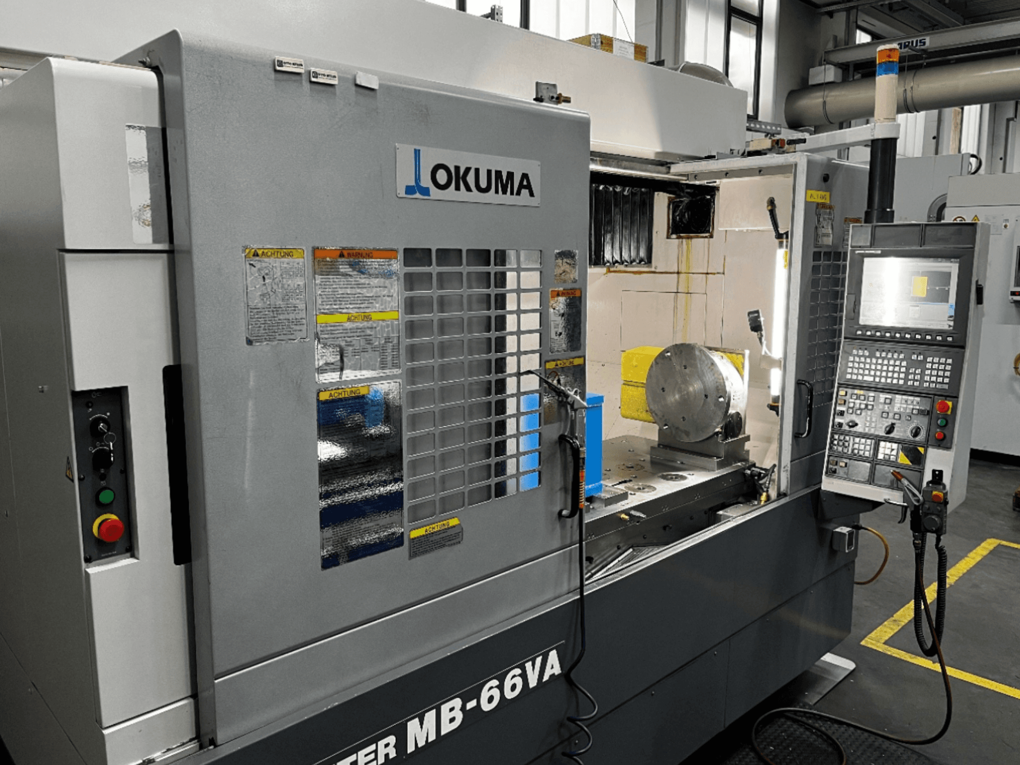 Вертикальний обробний центр Okuma MB66-VA спереду: панель керування, заготовка та внутрішня частина верстата з етикетками безпеки.