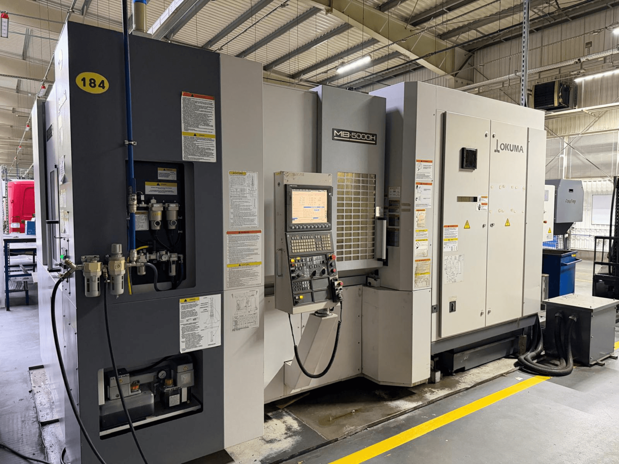 Вид спереду горизонтального обробного центру Okuma MB 5000H з елементами керування, маркуванням та гідравлічними з'єднаннями.