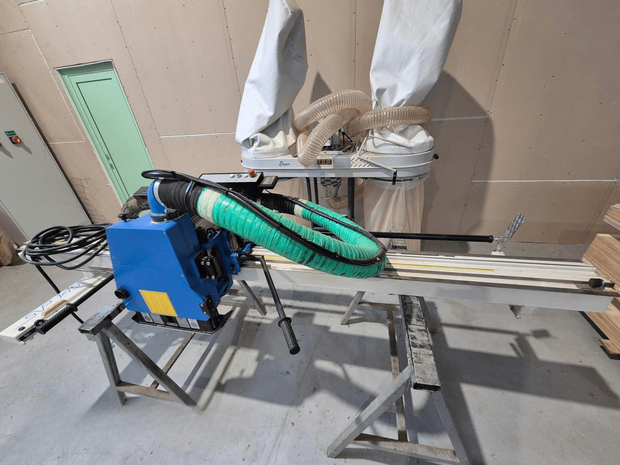 Горизонтальна панельна пила SVP-175 синього кольору із зеленим шлангом для пилу та підставкою, вид збоку в умовах майстерні.