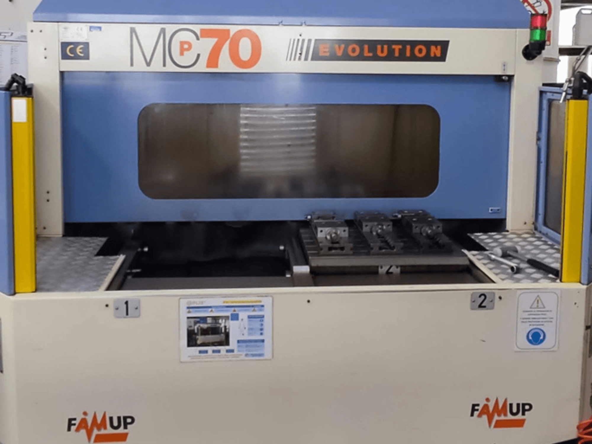 Вертикальний обробний центр FAMUP MCP70 E, вид спереду з панеллю керування та робочою зоною з кріпленнями та засобами безпеки.