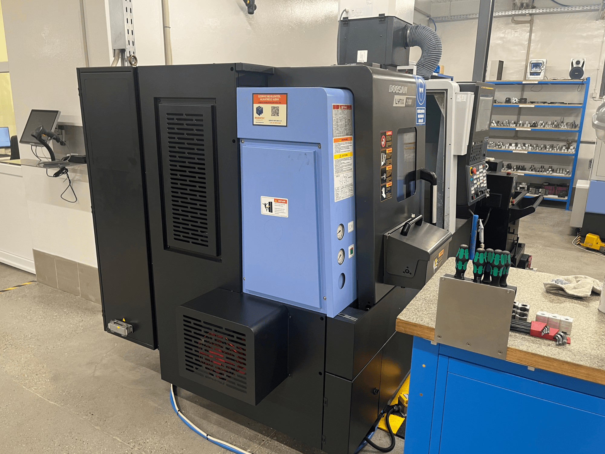 Токарний верстат Doosan Lynx 2100, вид збоку, демонструє панель управління, систему вентиляції та інструменти на робочому столі.