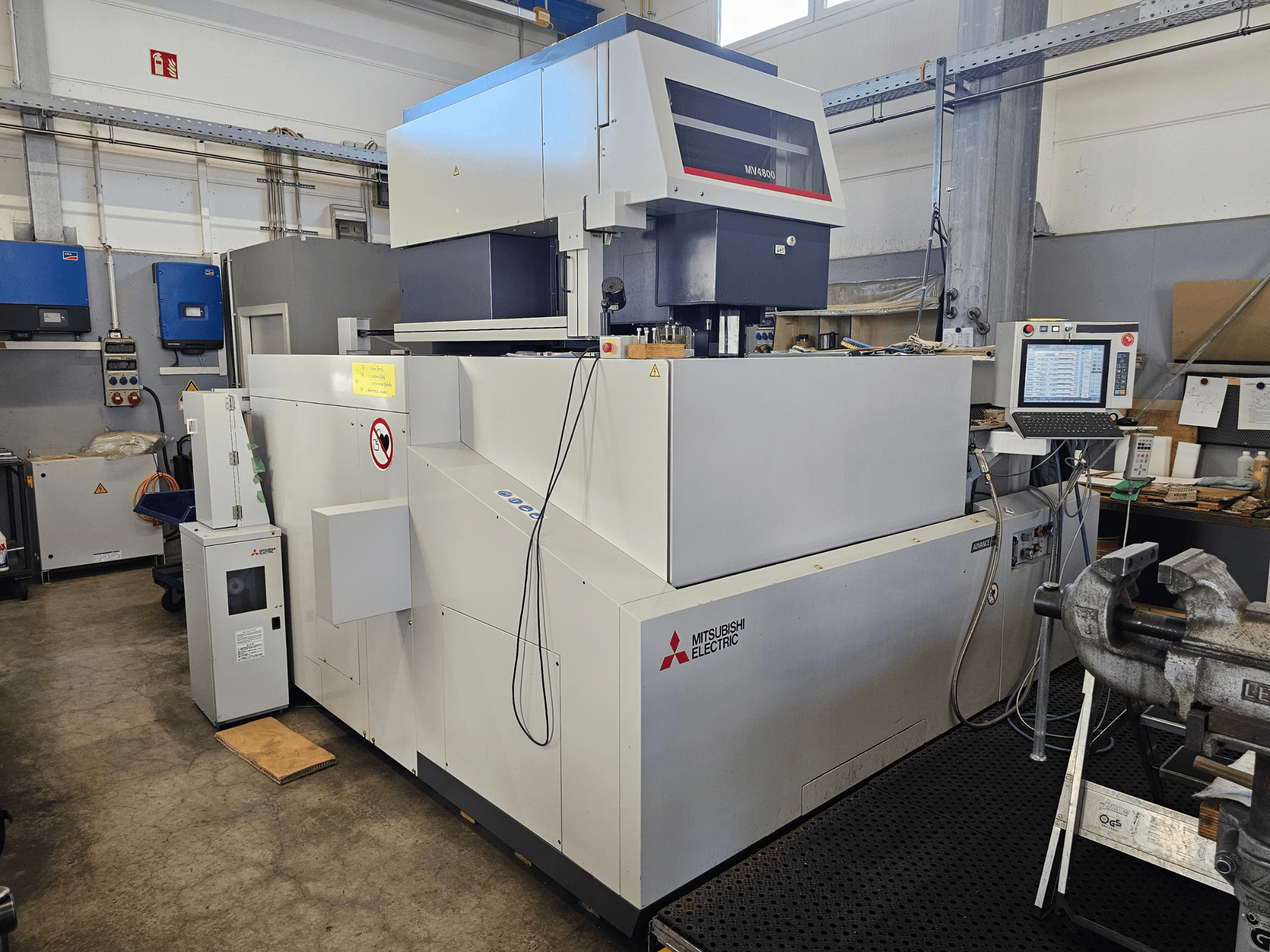 Дротяний електроерозійний верстат Mitsubishi MV4800 в майстерні, вид збоку: панель керування, етикетки безпеки та навколишнє обладнання.