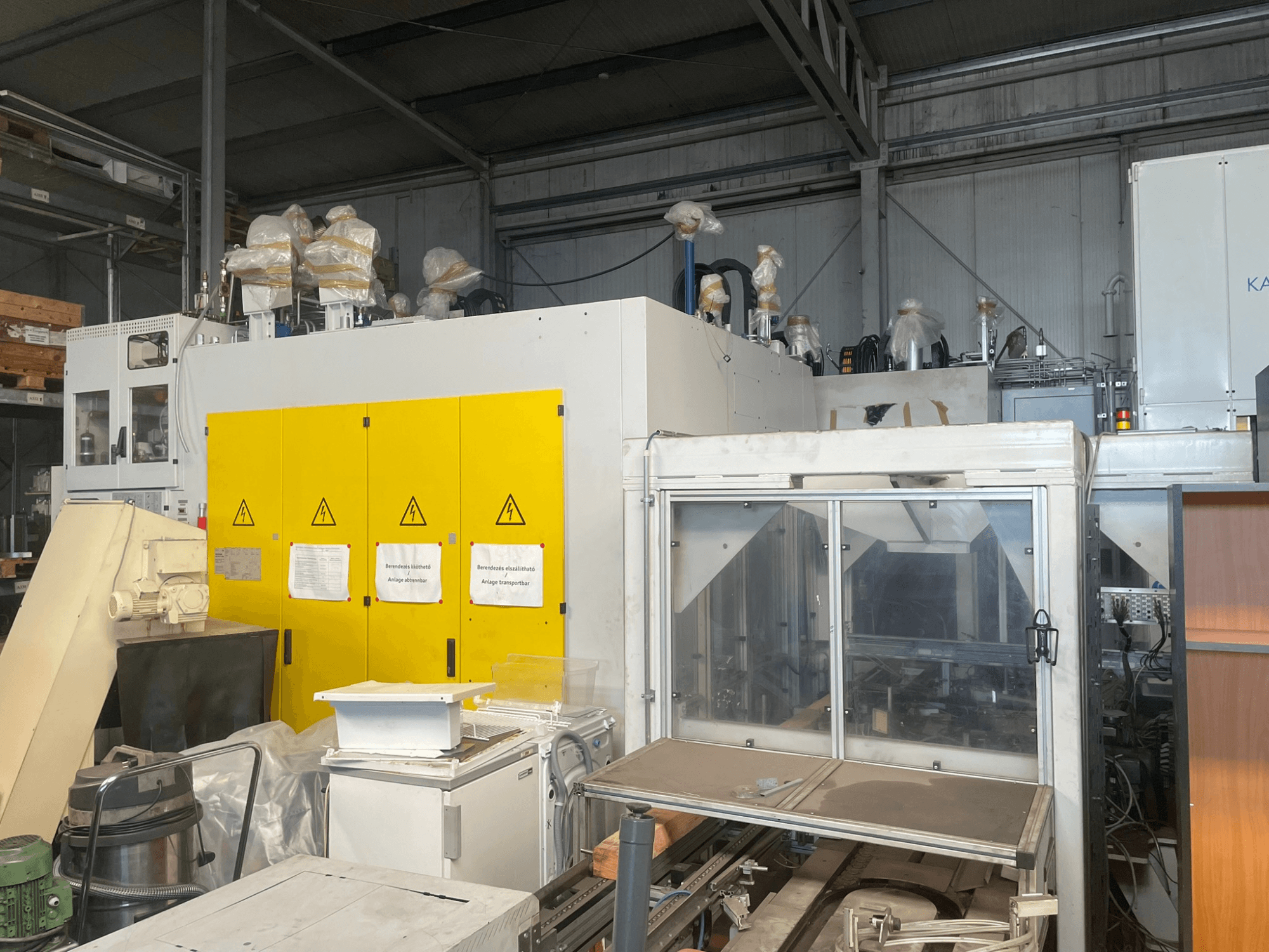 Промислове обладнання з жовтою панеллю керування, вид збоку, з попереджувальними етикетками та пластиковим покриттям, у складських умовах.