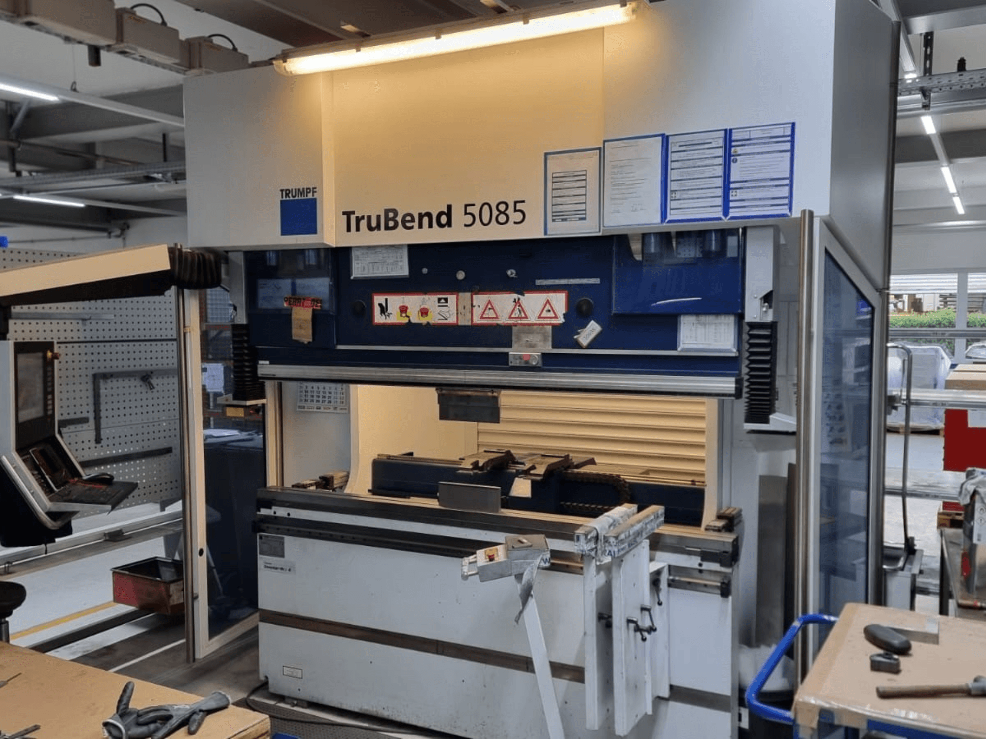 Згинальний верстат TRUMPF TruBend 5085, вид спереду, з панеллю керування, робочим місцем і попередженнями про безпеку в майстерні.