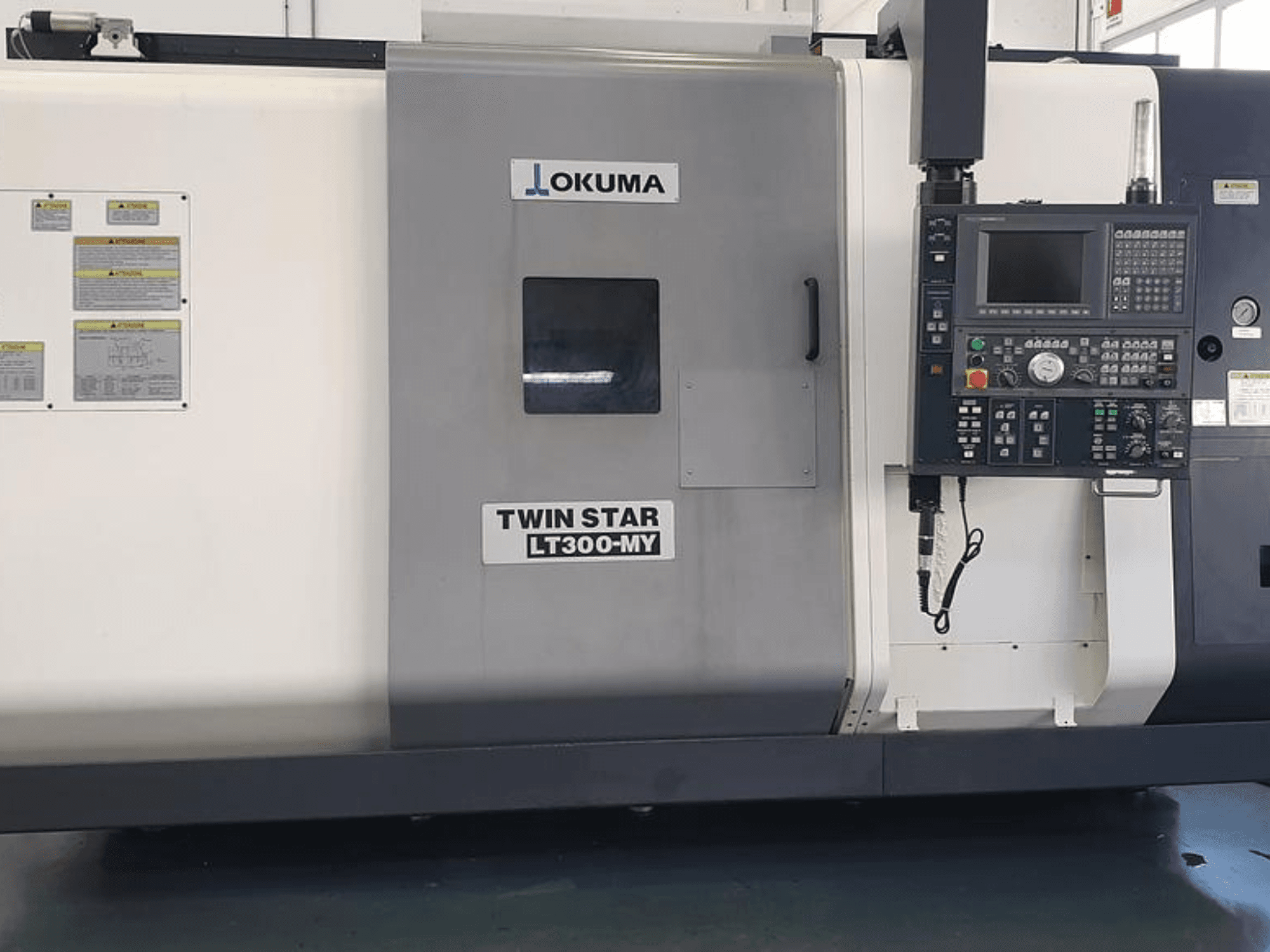 Токарний верстат з ЧПК Okuma Twin Star LT300-MY у вигляді збоку, з панеллю керування, кнопками керування та знаками безпеки.