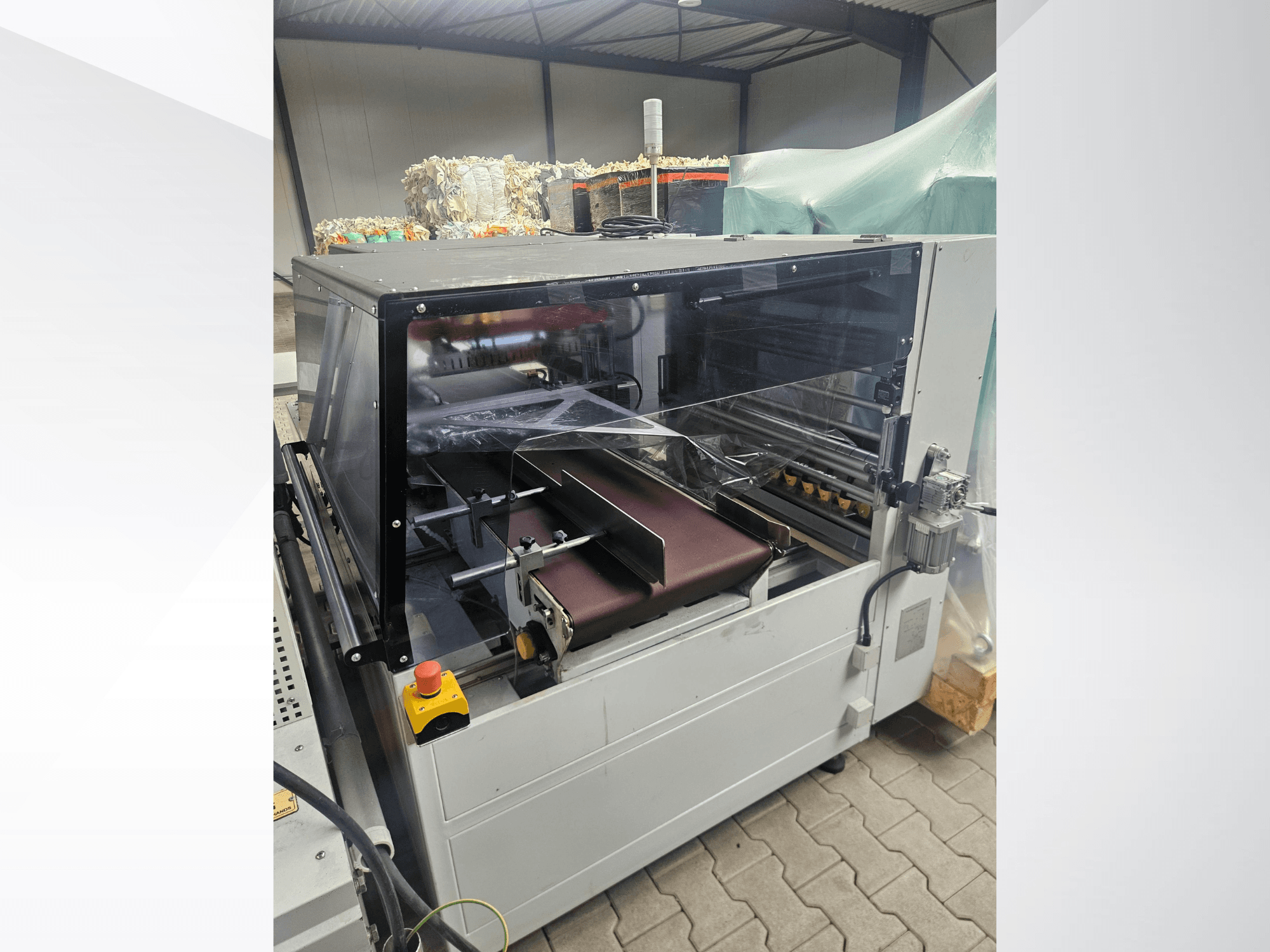 Машина AMTEC Speed Shrink 40 PRO, вид спереду, на якому видно механізм обробки пластику з прозорою захисною кришкою та конвеєрною стрічкою.