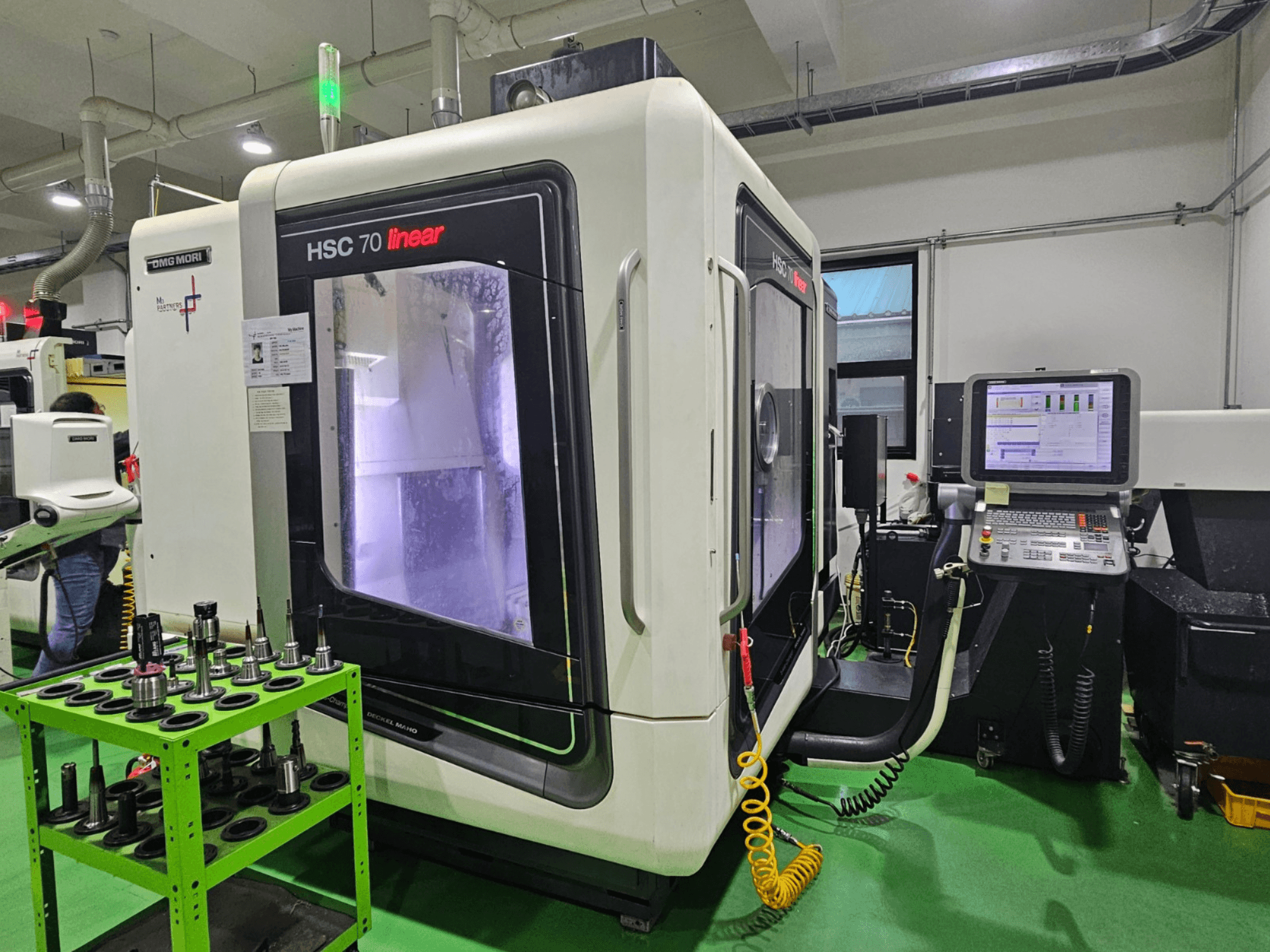 Лінійний універсальний обробний центр DMG MORI HSC 70, вид спереду, з тримачами інструментів на зеленому столі та панеллю керування.