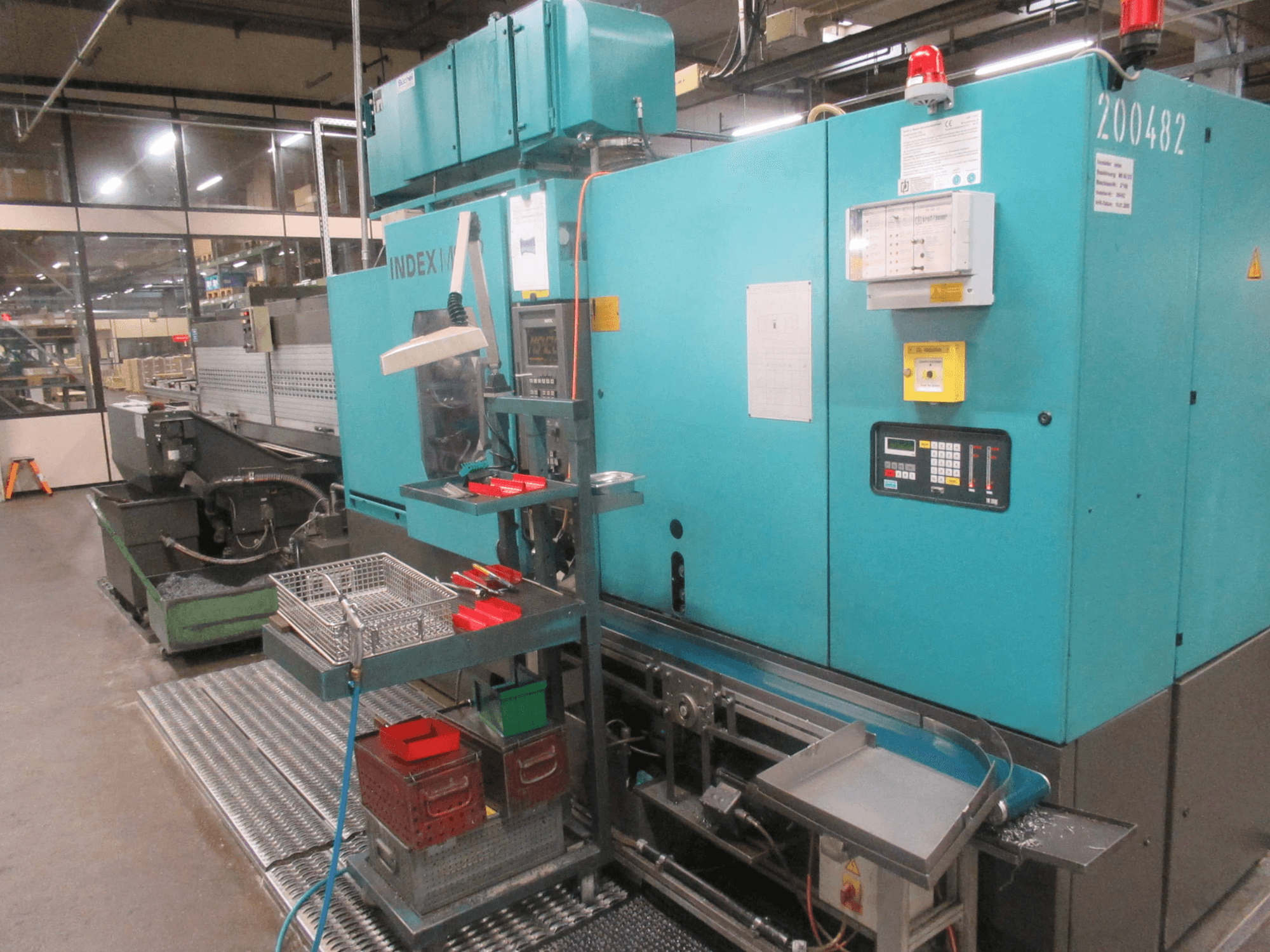 Бірюзовий верстат INDEX на заводі, вид збоку; має панель керування, сховище для інструментів і конвеєр. Видимі бункери, шланги та сигналізація.