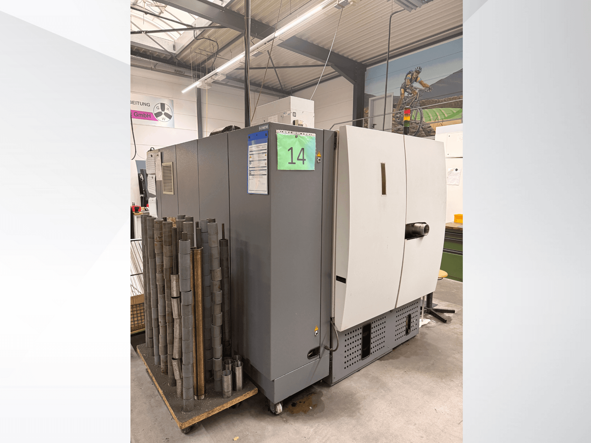 Сірий промисловий верстат від Siemens, вид збоку, з запасом циліндричних металевих деталей поруч та етикетками обладнання.