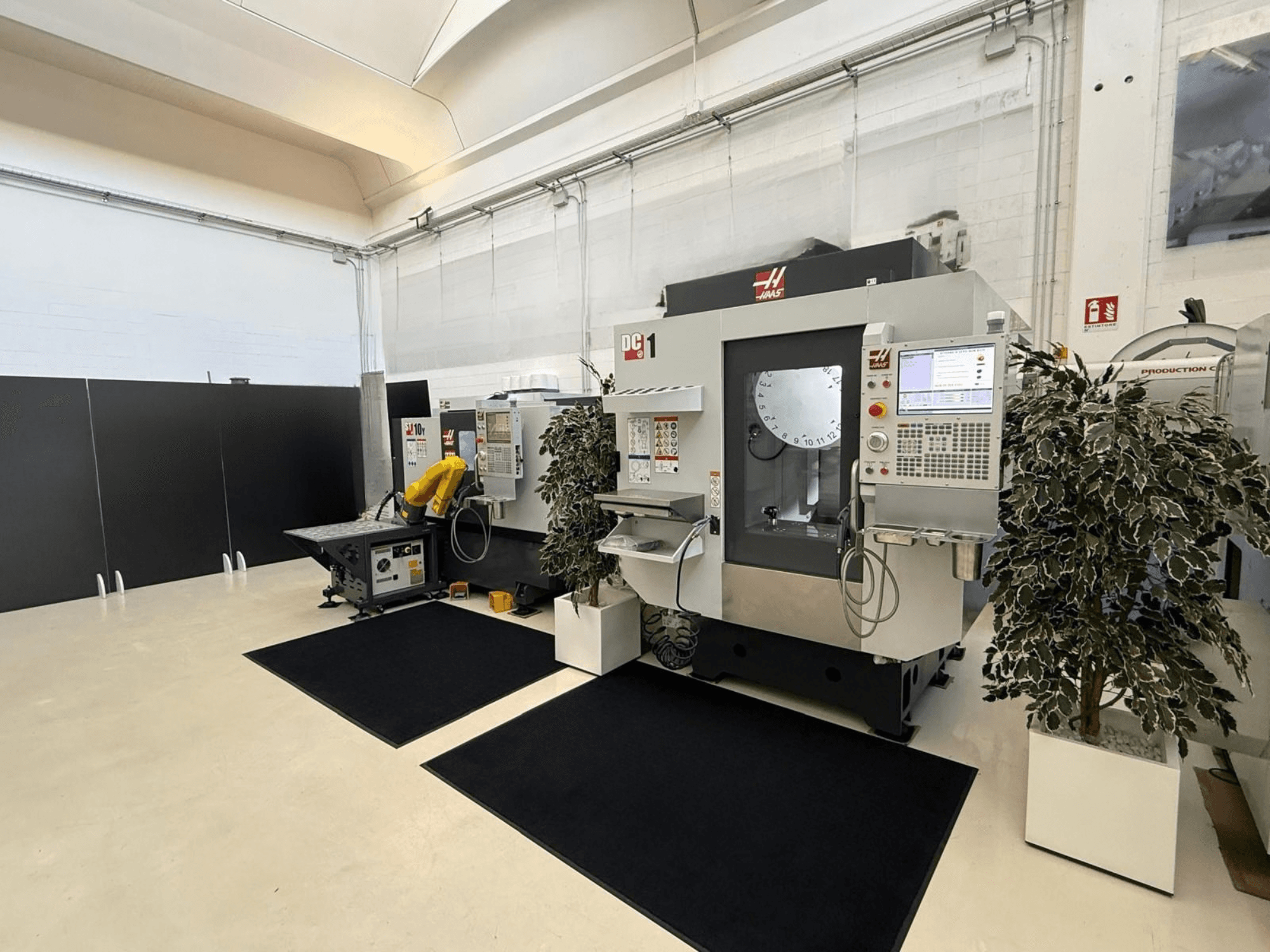 Вигляд спереду вертикального обробного центру HAAS DC-1 у чистому виробничому приміщенні, на якому видно сенсорну панель управління та обладнання, розташоване навколо.