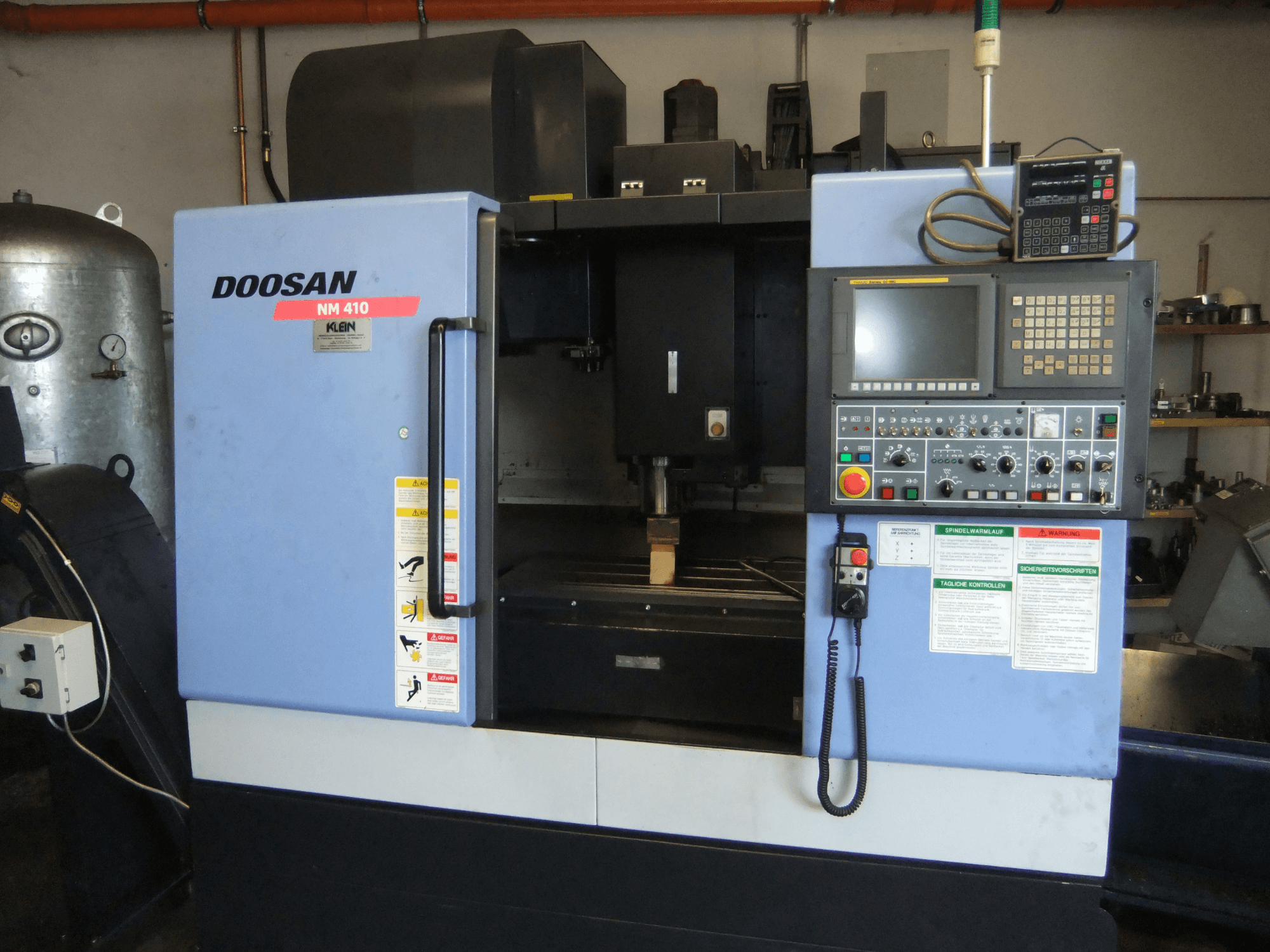 Вертикальний обробний центр DOOSAN NM 410, вид спереду, на якому видно панель управління, зону шпинделя та попереджувальні наклейки.