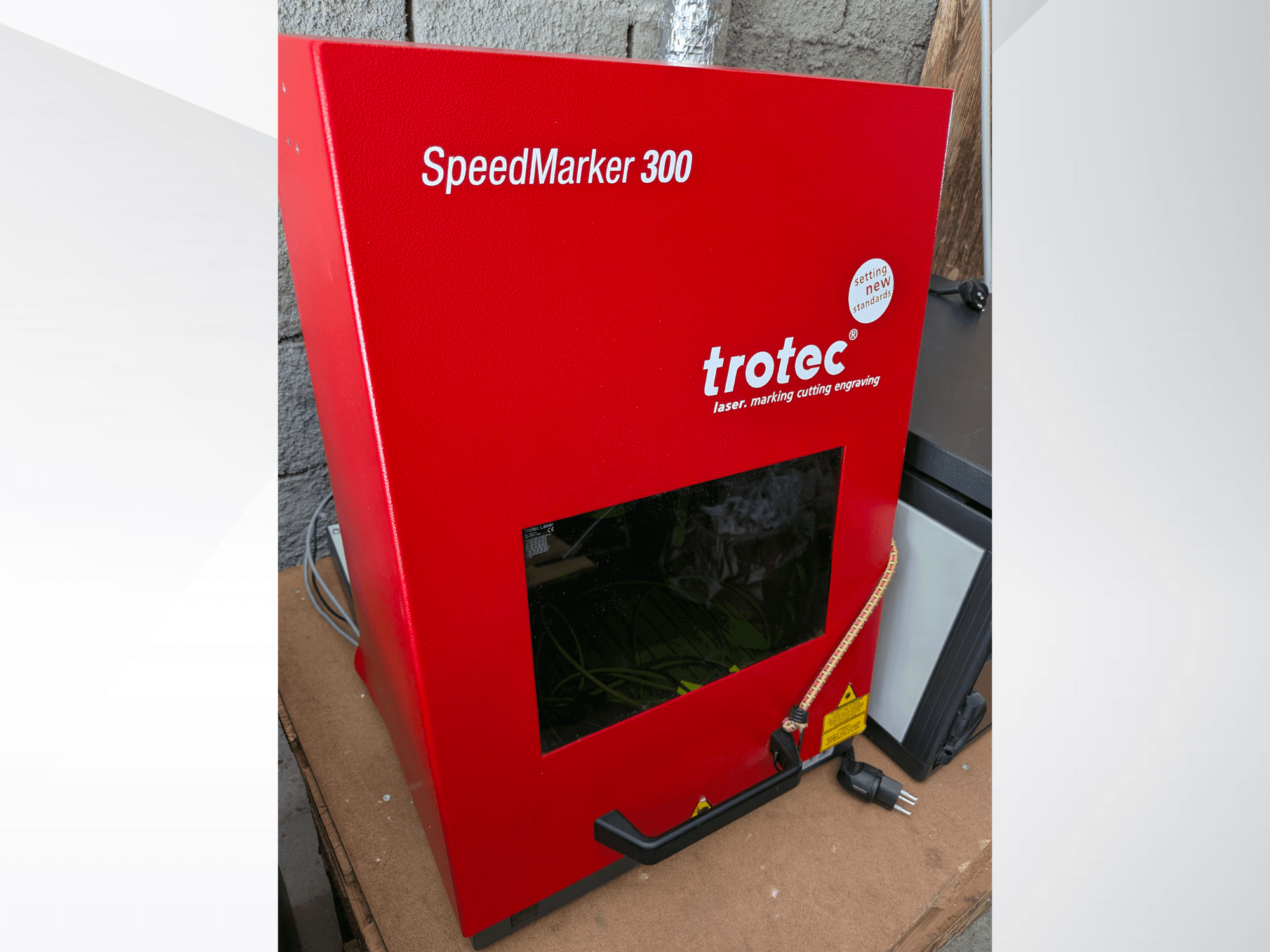 Червона лазерна маркувальна машина Trotec SpeedMarker 300, вид спереду, з великим оглядовим вікном і підключенням до електромережі.