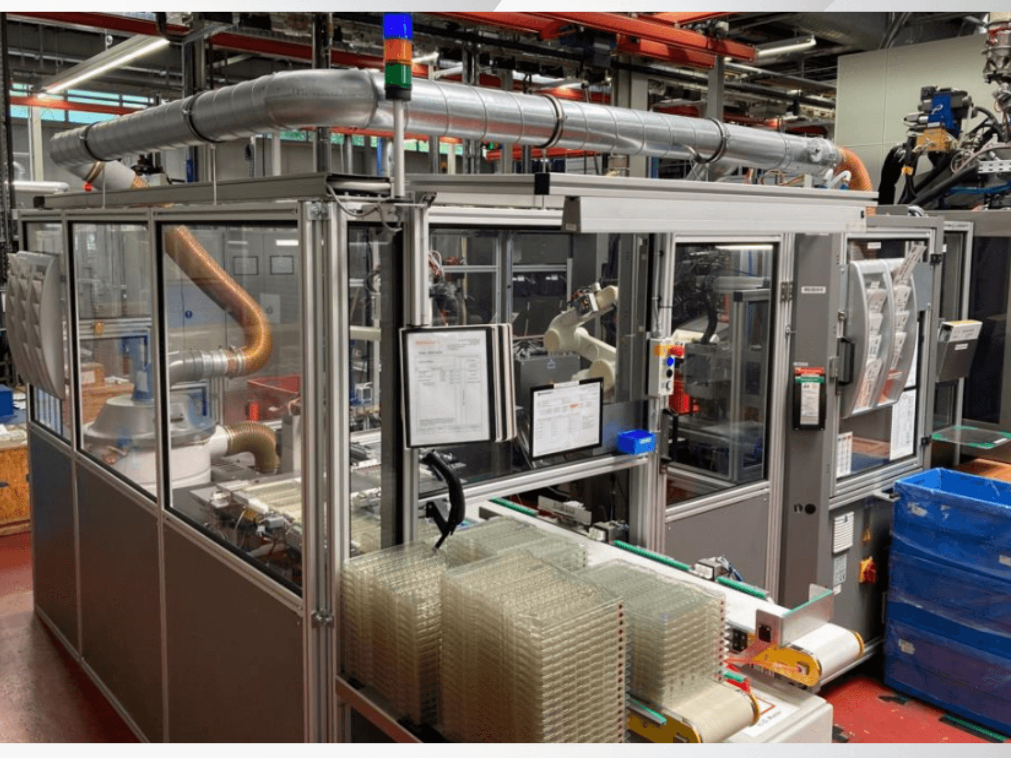 Вид спереду на автоматизовану машину з металевими трубами, панеллю керування та складеними в штабелі прозорими пластиковими контейнерами на заводському майданчику.