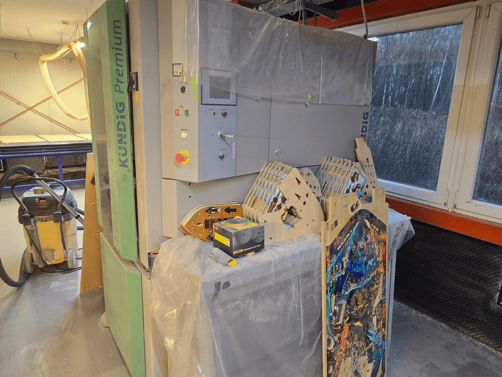 Стрічкова шліфувальна машина Kundig Premium-1 1100-C в майстерні, з панеллю керування та заготовками з деревини, вид збоку, з пилососом на задньому плані.