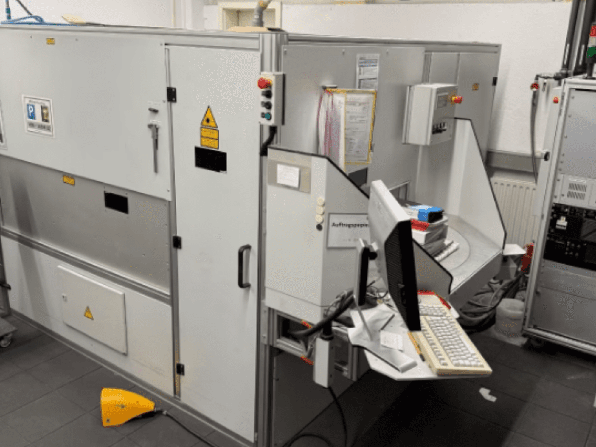 Промислова машина з комп'ютерним терміналом, вид збоку; включає кнопки керування та попереджувальні знаки безпеки.