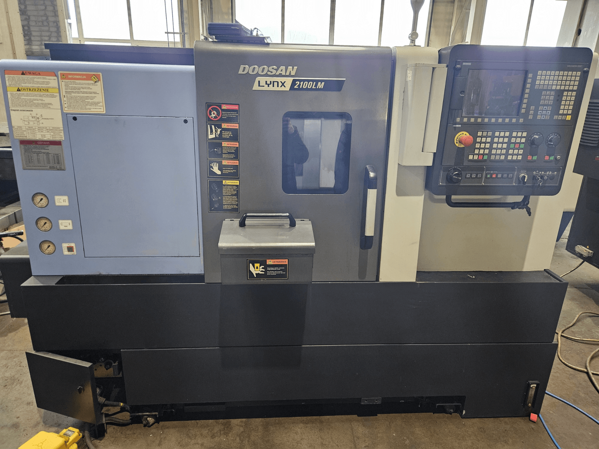 Токарний верстат з ЧПУ Doosan Lynx 2100LM, вид спереду, з панеллю керування, манометрами та етикетками безпеки.