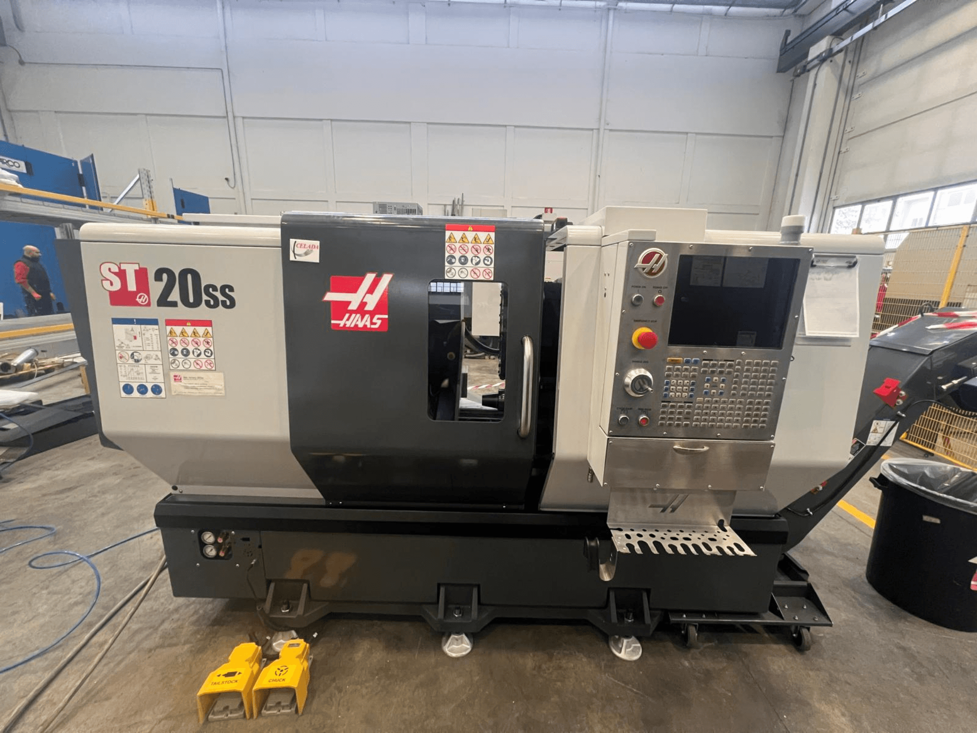Токарний верстат з ЧПК Haas ST 20SS у вигляді збоку, з панеллю керування, знаками безпеки та ножними педалями на підлозі.
