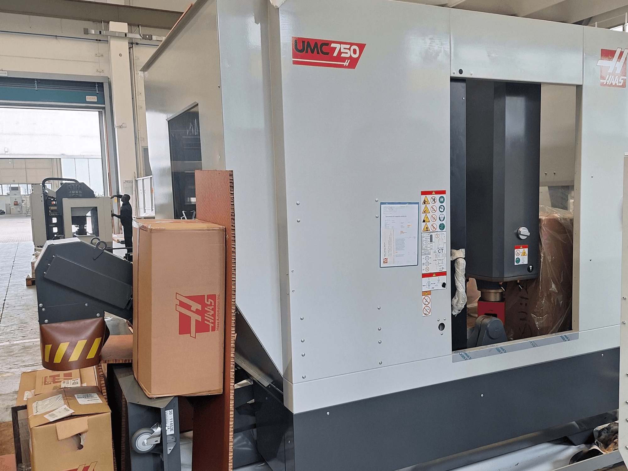 Універсальний обробний центр HAAS UMC-750, вид збоку, на якому показано панель керування та відсік для зберігання інструментів із попереджувальними наклейками та в упаковці.