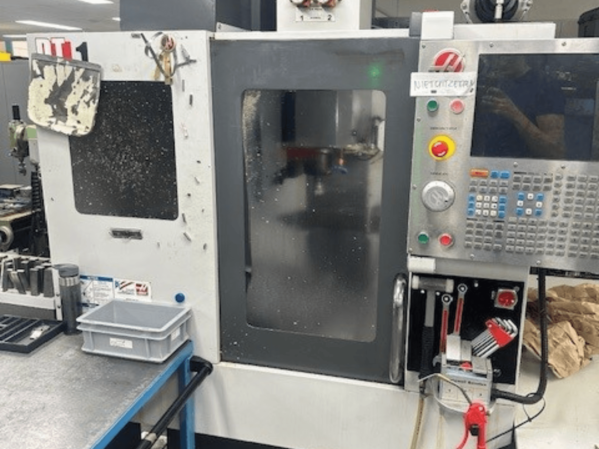 Вертикальний обробний центр Haas DT-1, вид спереду з панеллю керування, видимими забрудненнями та лотком для інструментів на робочому столі.