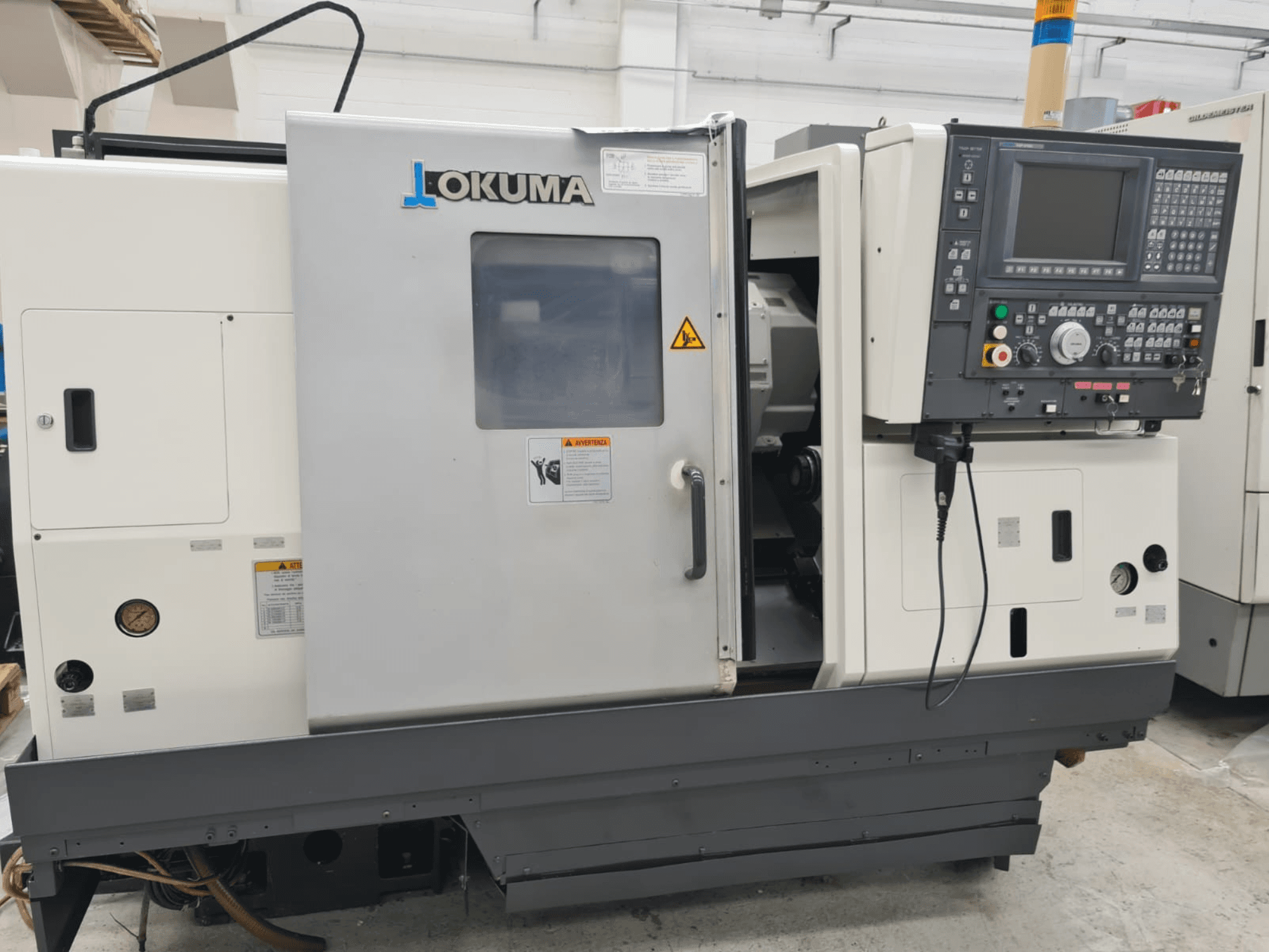Горизонтальний токарний верстат Okuma LB 300M X 500 у вигляді збоку, демонструє панель керування, доступ до шпинделя та попередження з техніки безпеки.