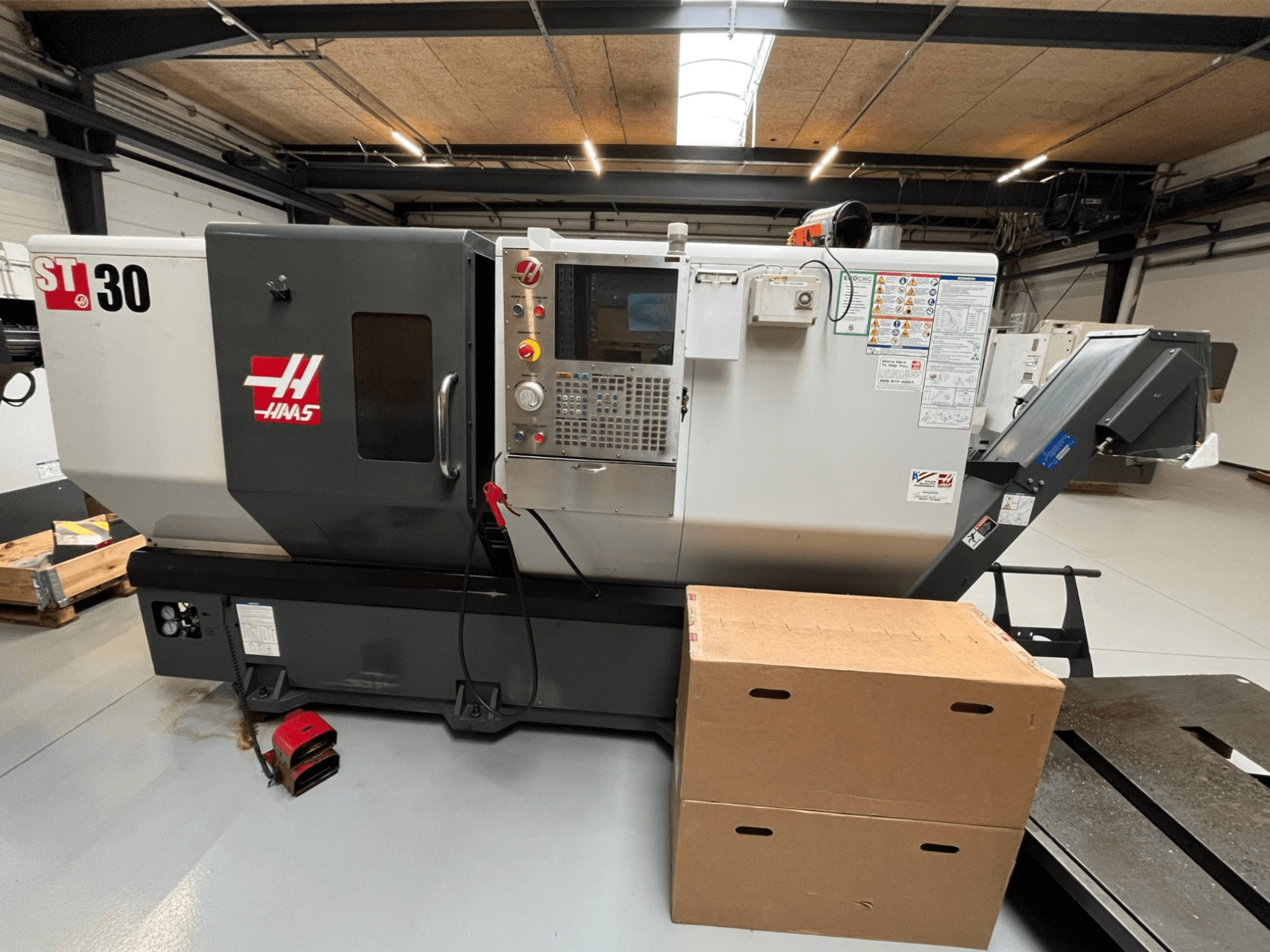 Токарний верстат з ЧПК Haas ST-30, вид спереду, має панель керування з кнопками, аварійну зупинку та дві складені картонні коробки.