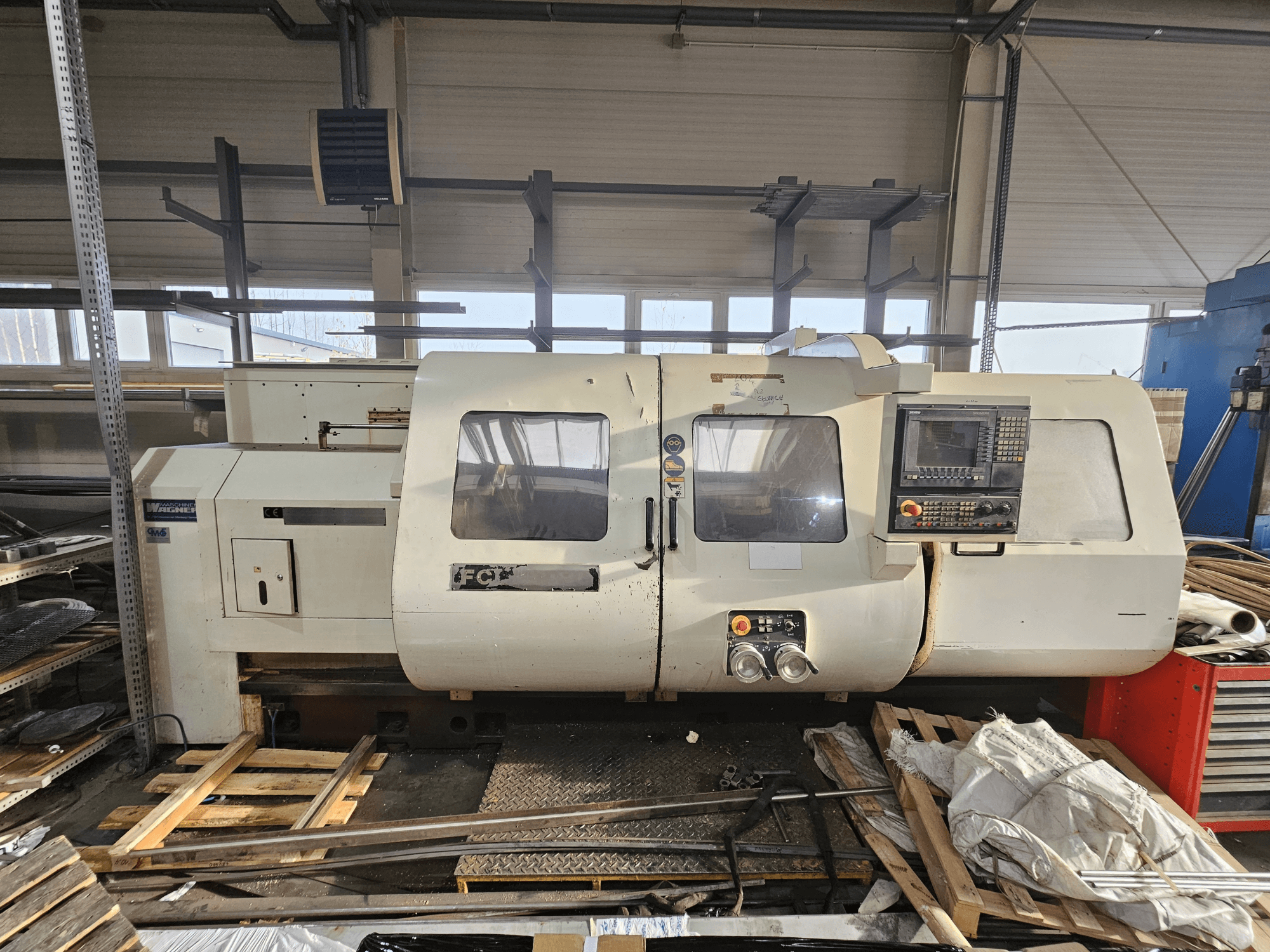 Токарний верстат з ЧПК Wabeco, вид збоку, з панеллю керування, вікнами та робочою зоною; обладнання на дерев'яних піддонах в умовах майстерні.