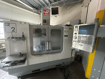 Верстат з ЧПК Haas VF-2, вид спереду, з елементами керування, тримачем інструменту та прозорими дверцятами з видимим інструментарієм всередині шафи.