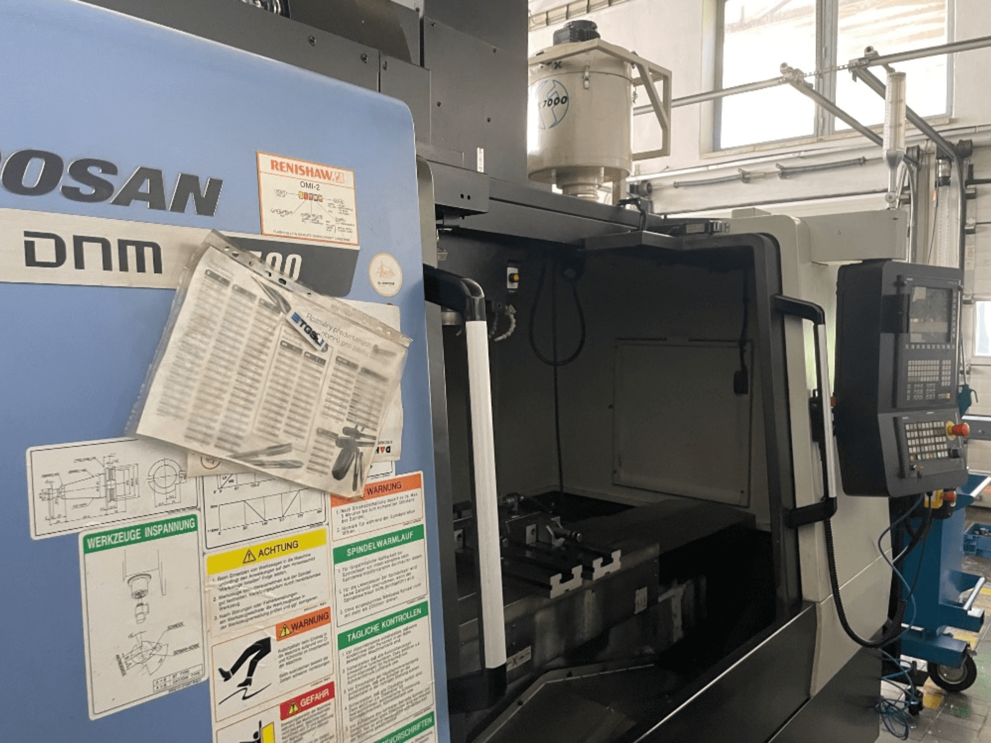Вертикальний обробний центр DOOSAN DNM 5700, вид спереду, показано панель керування, етикетки безпеки та деталі верстата.