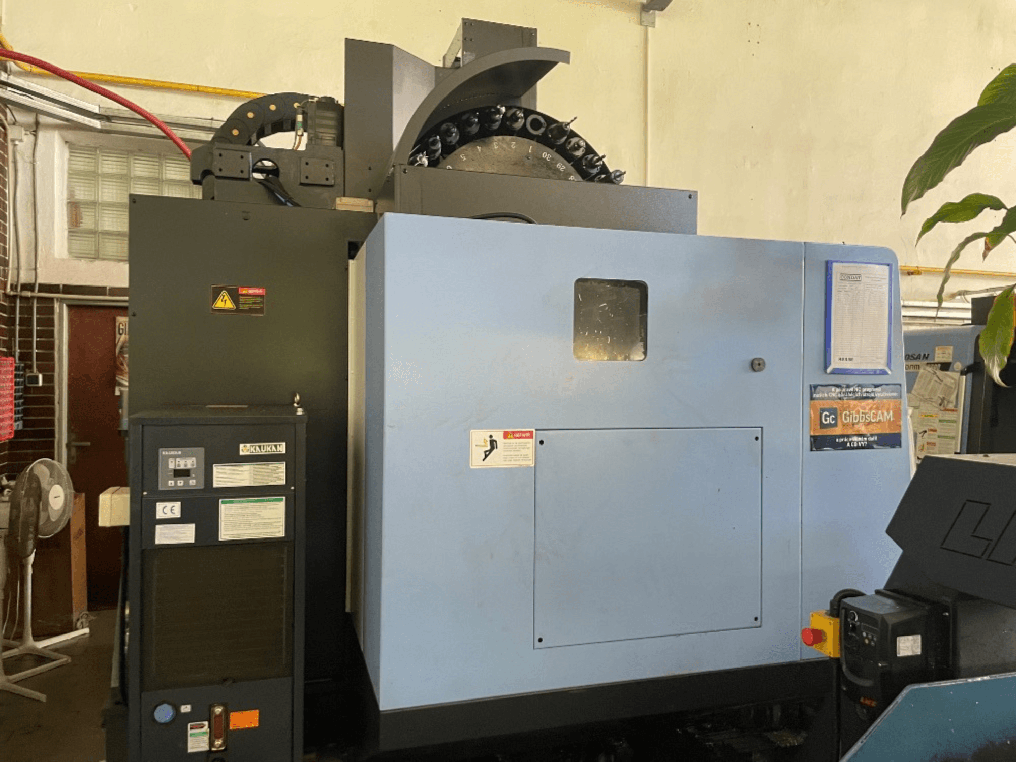 Вид спереду вертикального обробного центру DOOSAN DNM 5700 з панеллю керування, етикетками безпеки та функціями верстата.