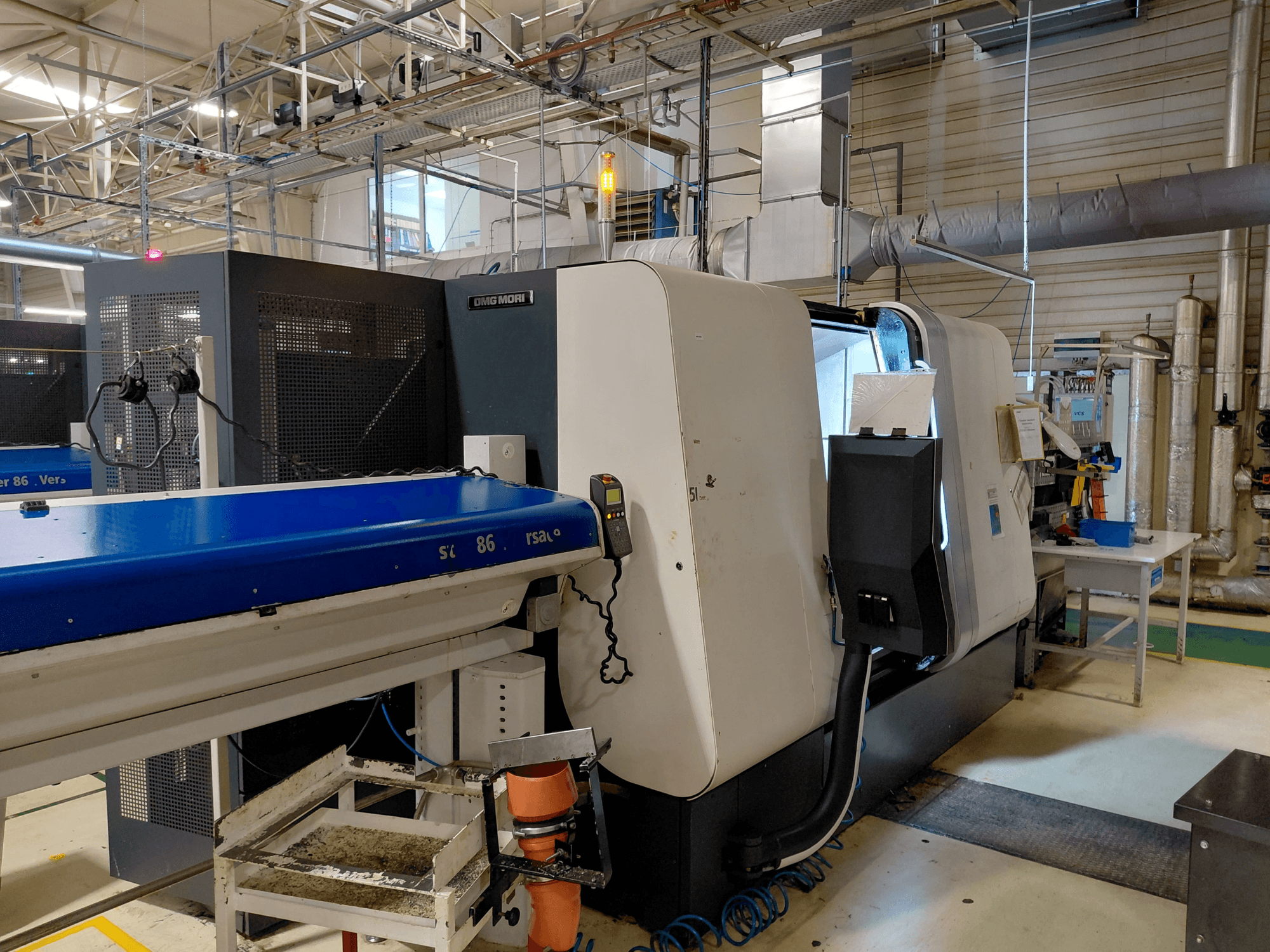 Лінійний багатошпиндельний токарний верстат DMG MORI Sprint 65, вид збоку з панеллю керування та конвеєром в умовах майстерні.