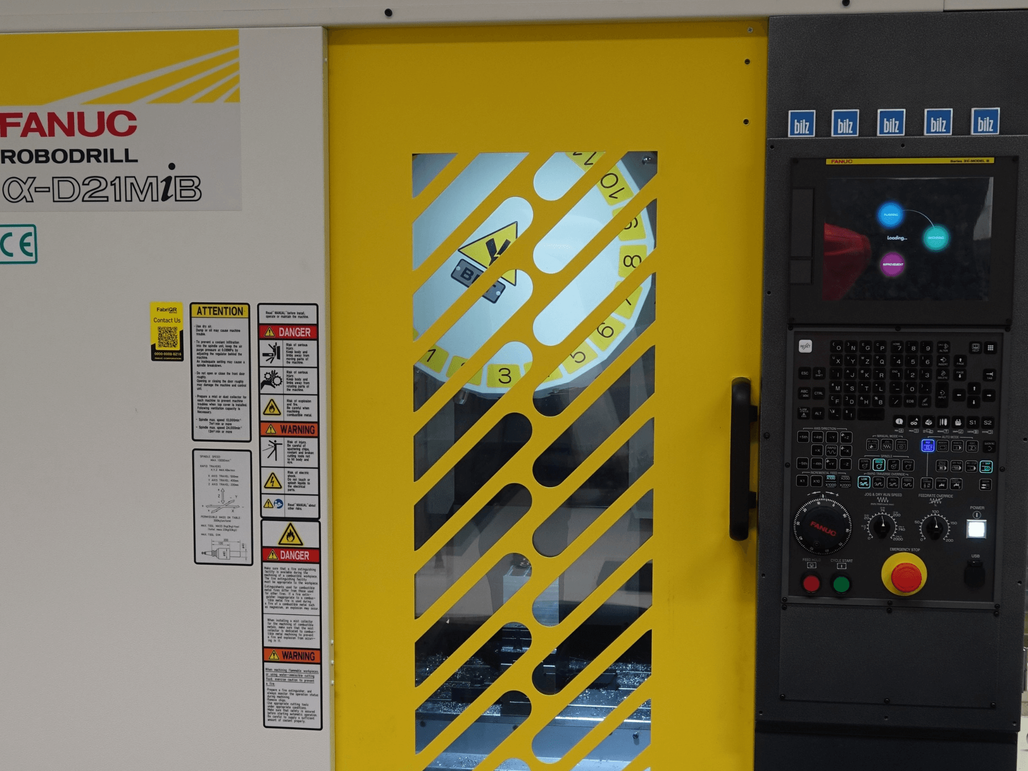 Вертикальний обробний центр FANUC α-D21MiB у фронтальному ракурсі: на зображенні видно попереджувальні написи щодо безпеки, панель управління та жовті захисні дверцята.