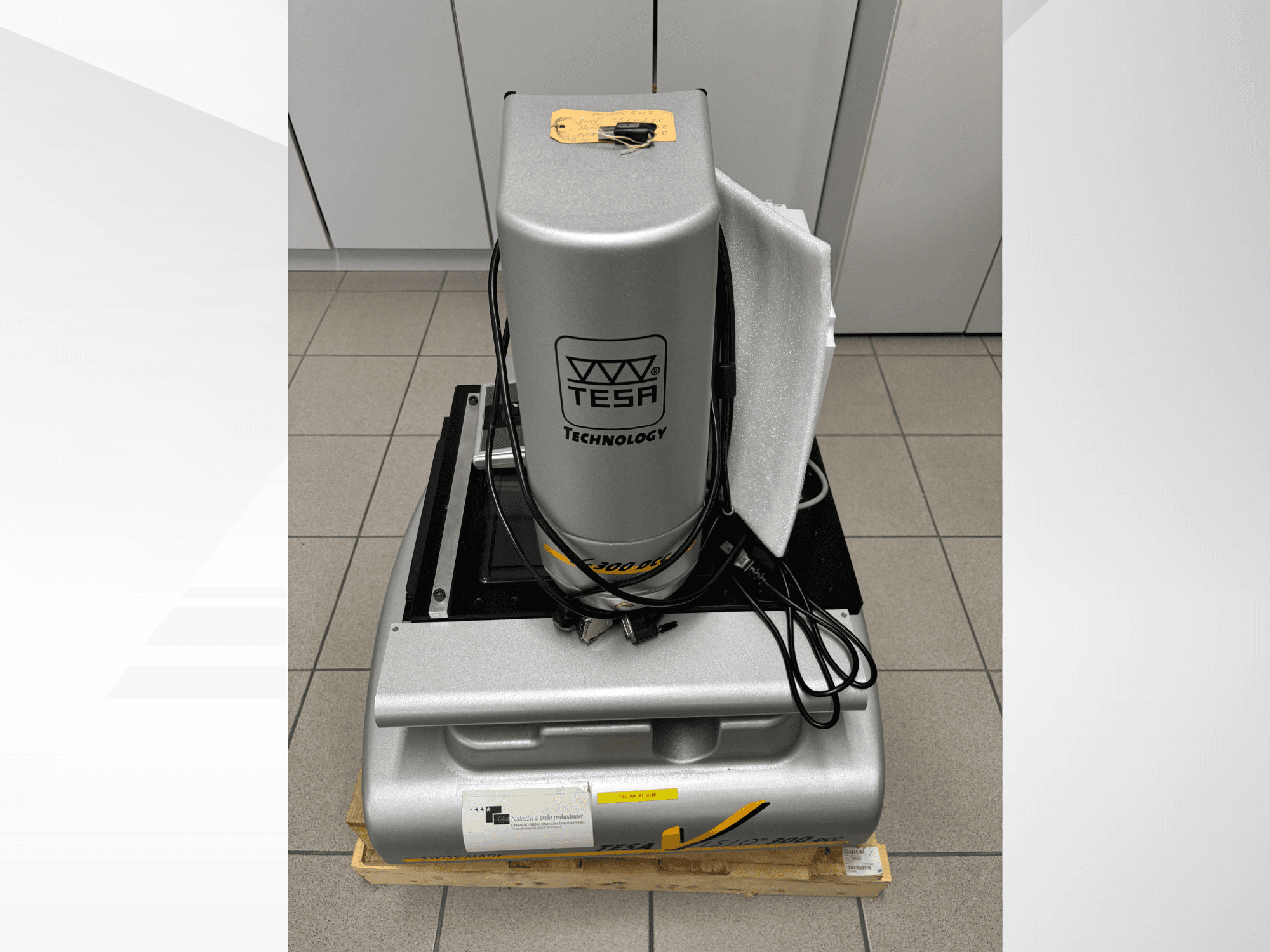 Оптична вимірювальна машина TESA Visio 300 DCC спереду з кабелями, на дерев'яному піддоні, видно захисну упаковку.