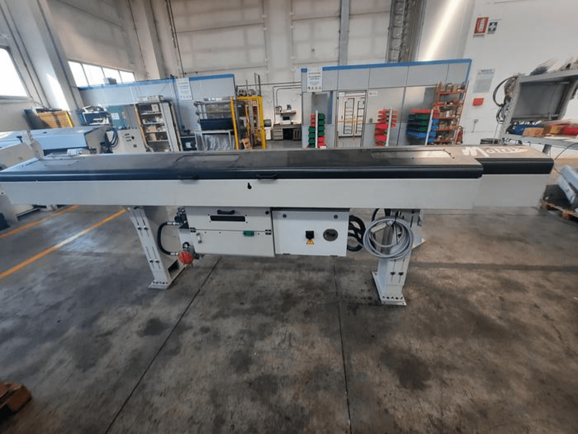 Металевий конвеєрний стіл з гладкою чорною стільницею, вид збоку, з кнопками керування та кабелями; частково видно логотип бренду.