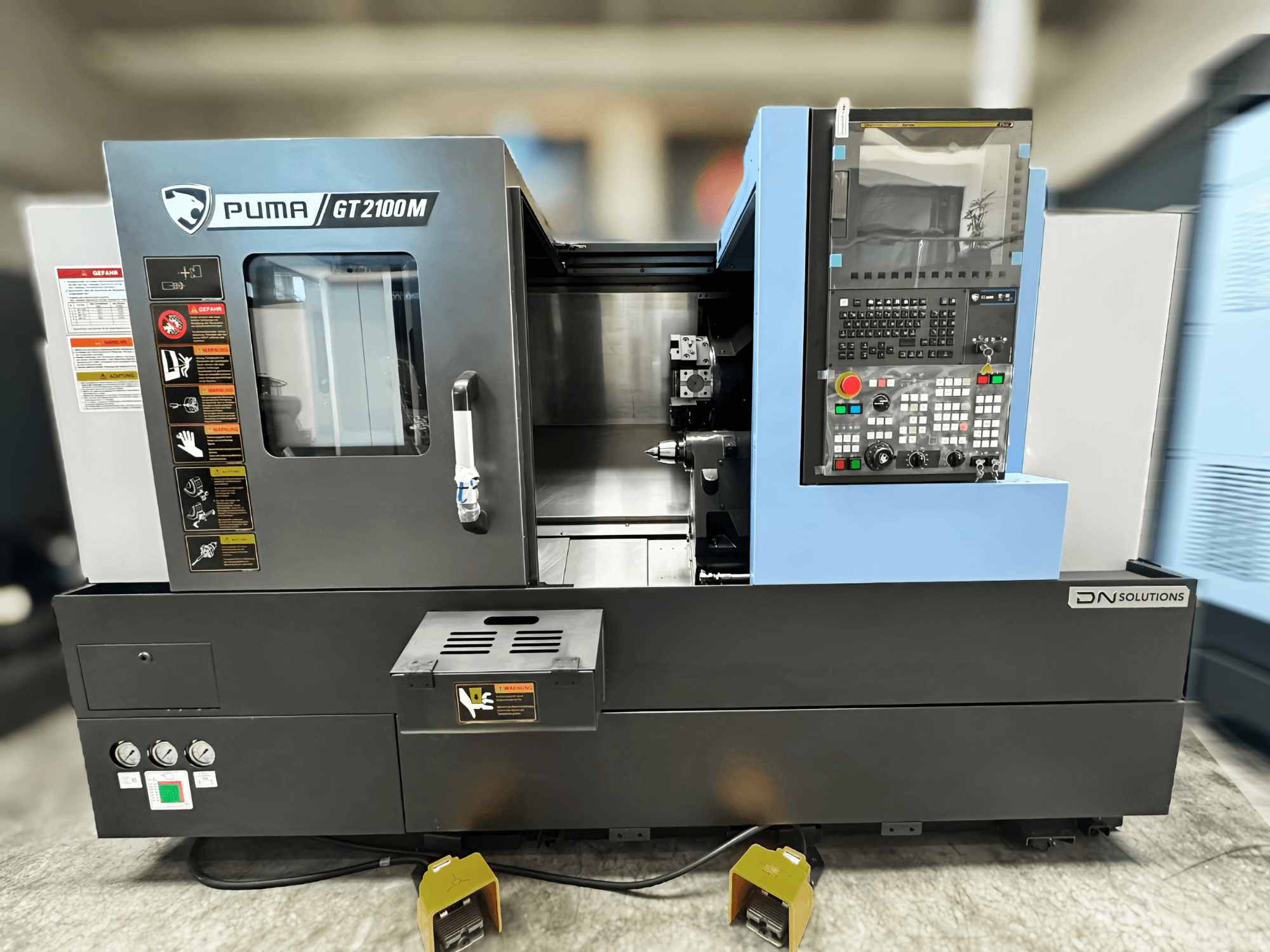 Токарно-фрезерний центр DN SOLUTIONS PUMA GT2100M, вид спереду, на якому видно панель керування, обробну камеру та знаки безпеки.