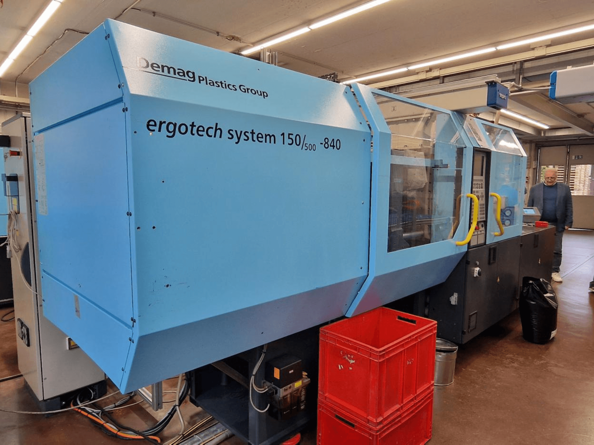 Термопластавтомат Demag Ergotech System 150/500-840 синього кольору, вид збоку, з ящиками та оператором на задньому плані.