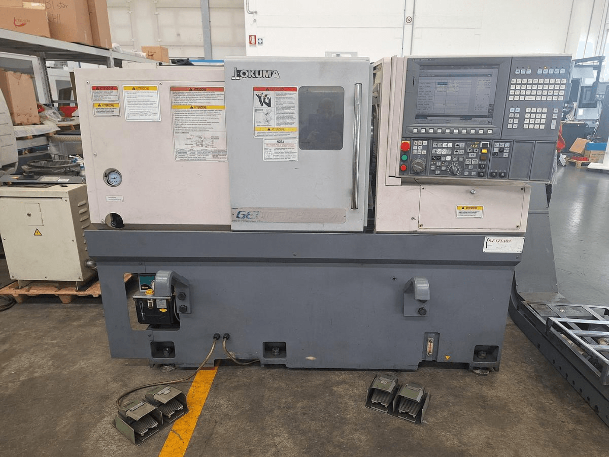 Горизонтальний токарний верстат Okuma GENOS L200E-M у вигляді збоку; панель керування, попередження про безпеку та ножні педалі.