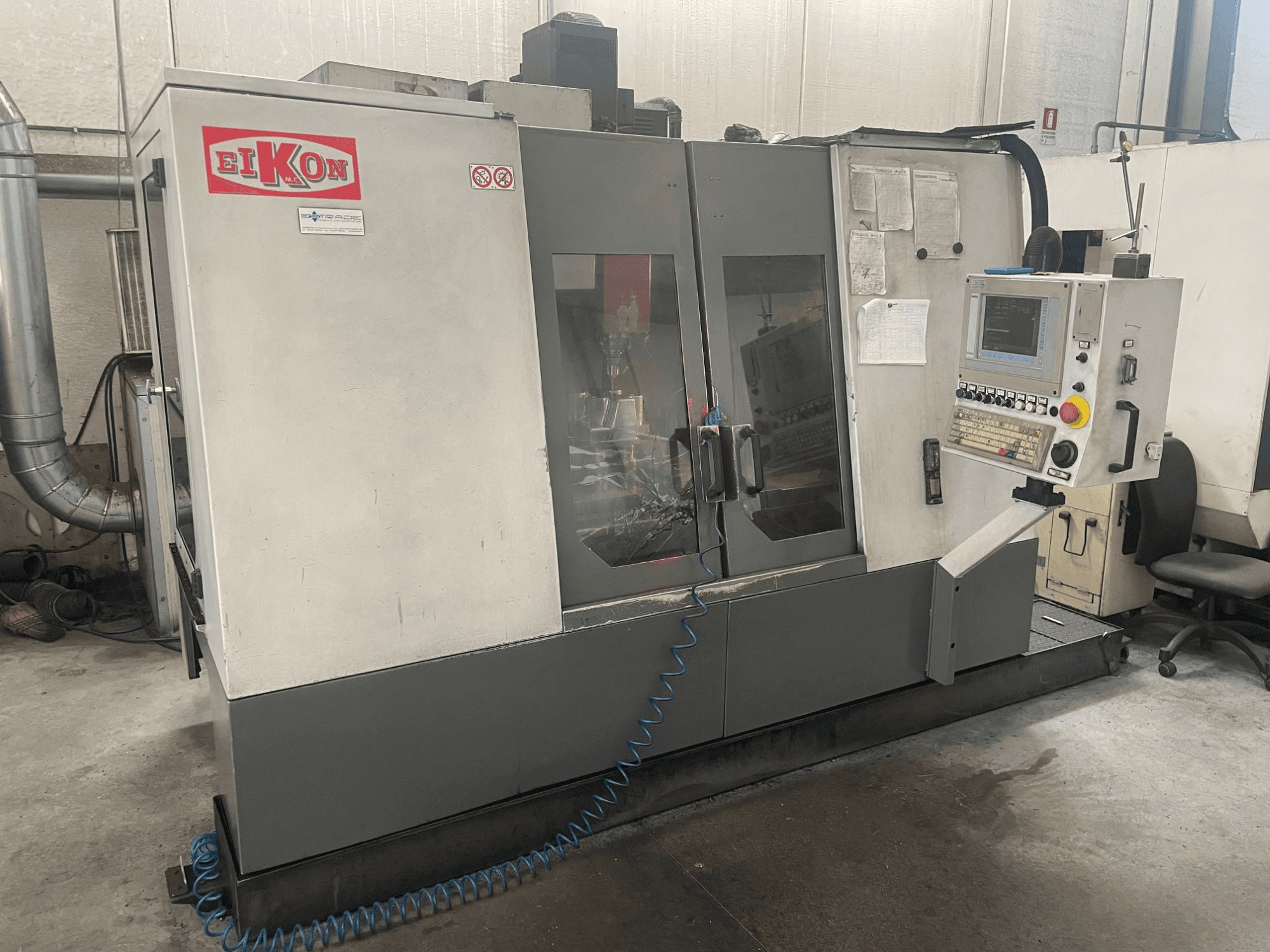 Верстат з ЧПУ EIKON, вид спереду, з панеллю керування, великими скляними дверима та синім шлангом для подачі повітря на підлозі.