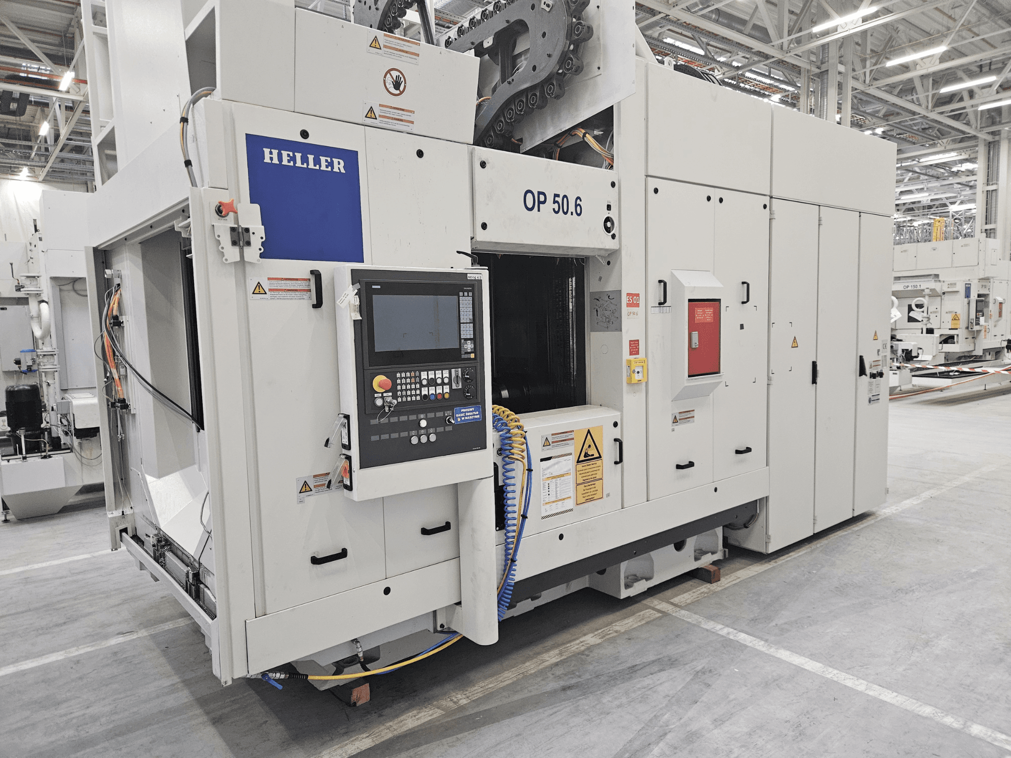 Обробний центр Heller OP 50.6, вид спереду, з панеллю керування, попереджувальними табличками та шлангами, видимими у заводському стані.