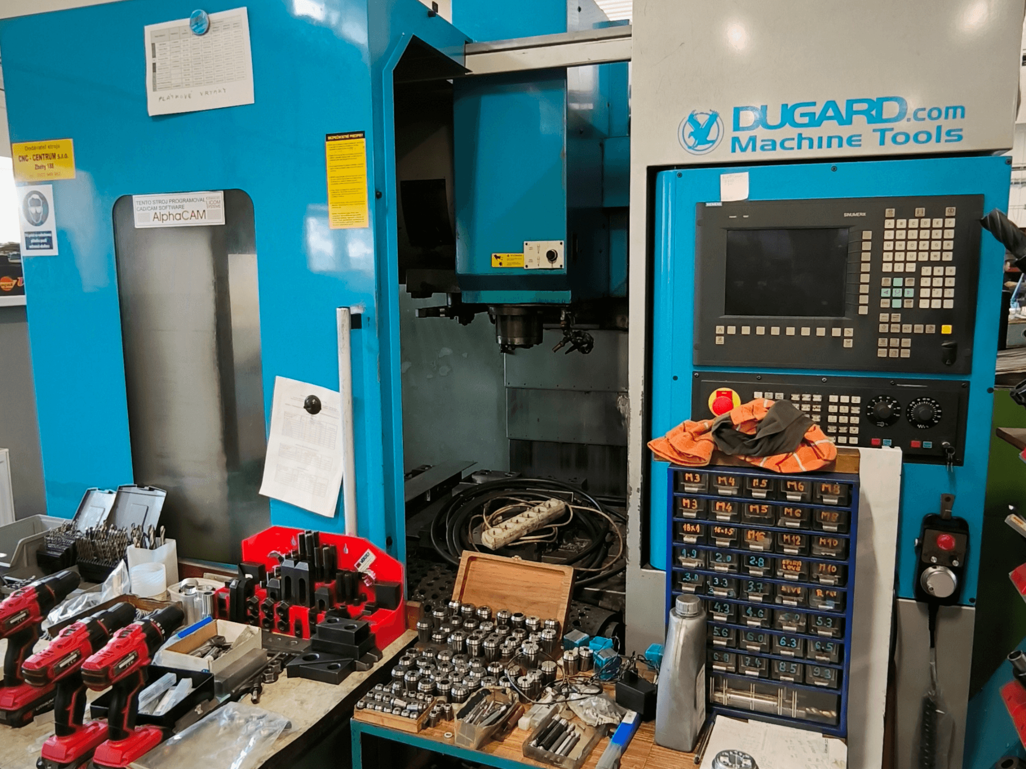 Вид спереду вертикального обробного центру Dugard 850VMC з панеллю керування, інструментами та аксесуарами на робочому столі.