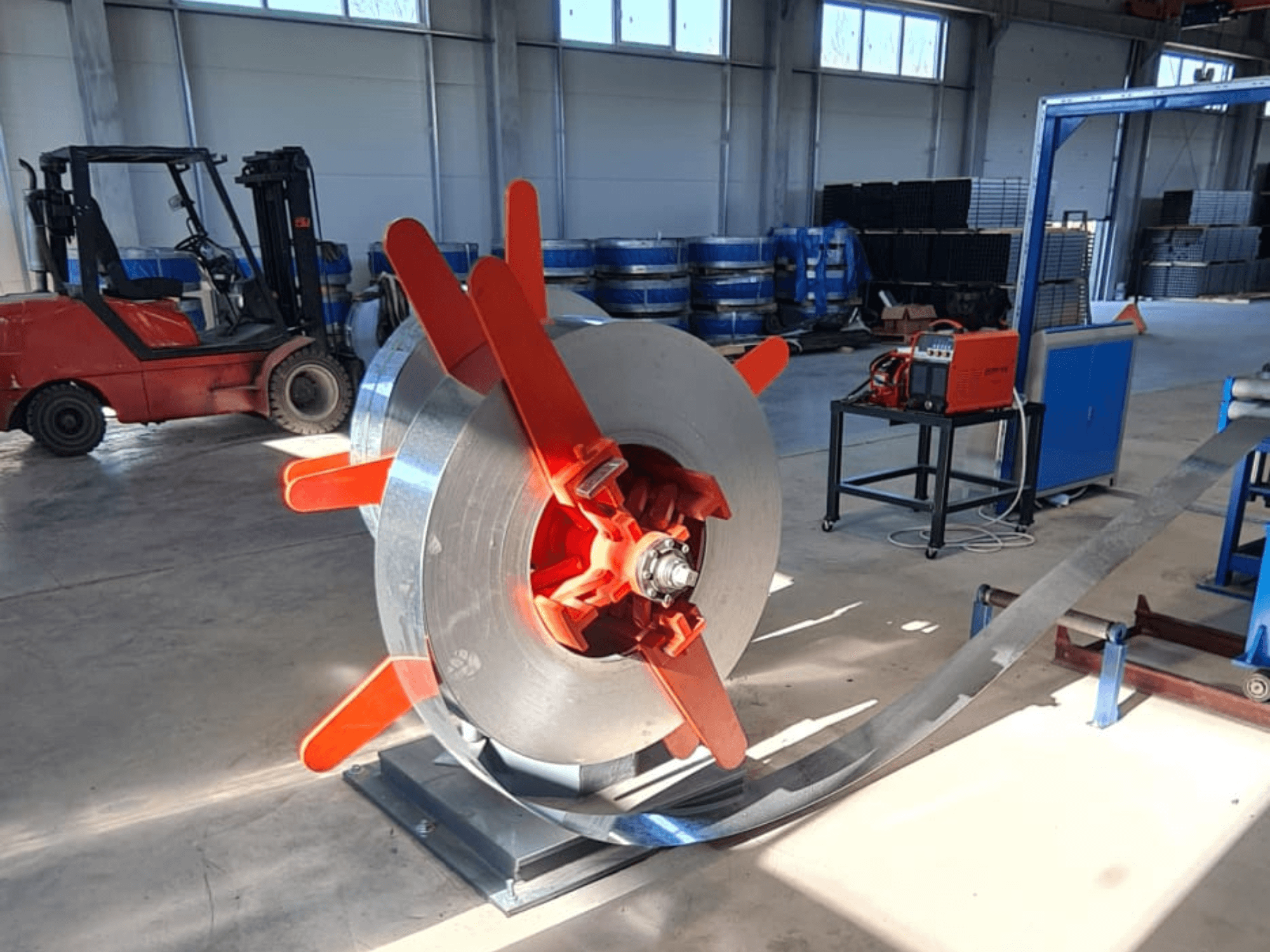 Металева котушка з червоними ручками на підставці, вид під кутом. Індустріальне тло з машинами та навантажувачем.