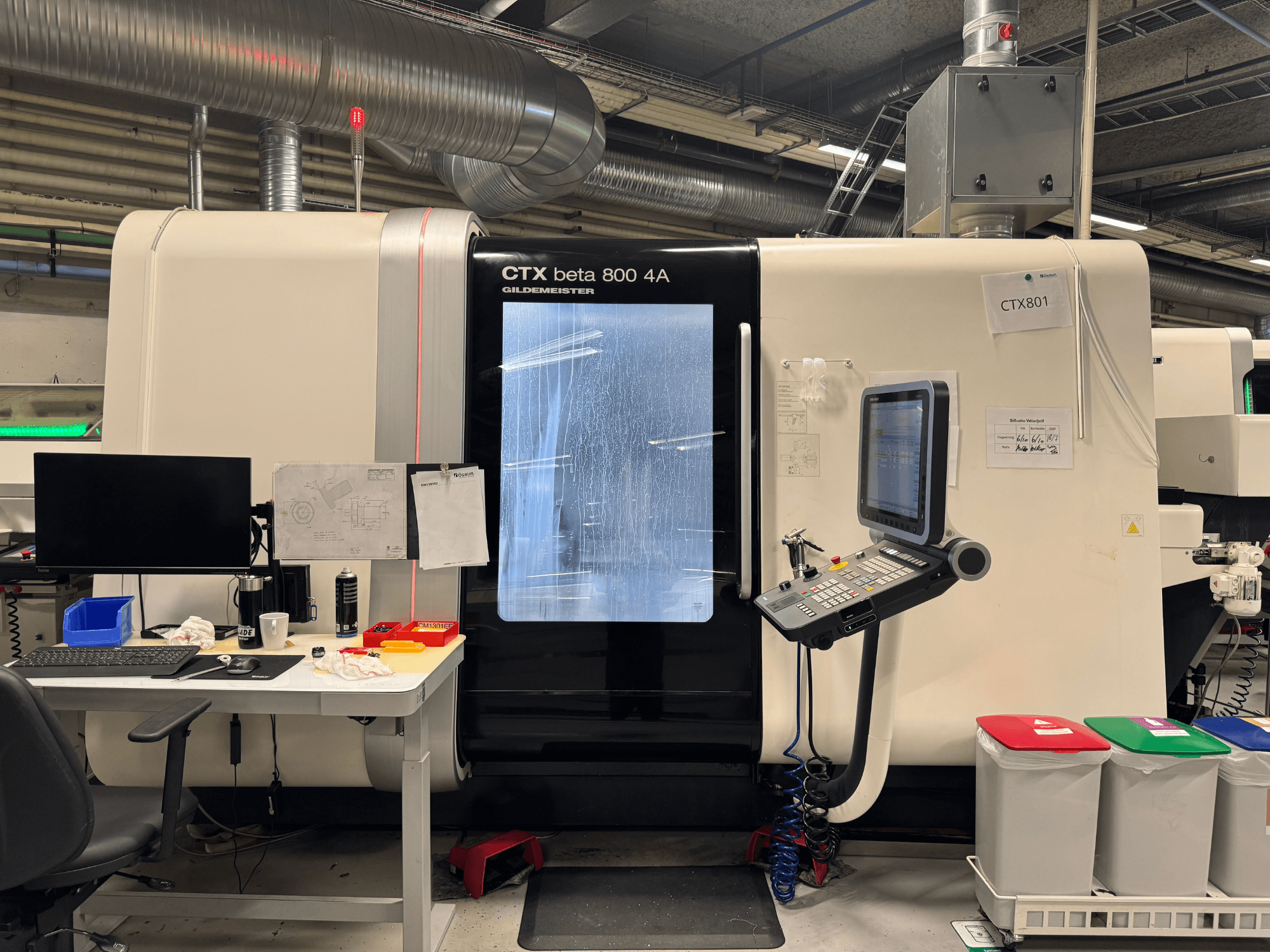 Верстат CTX beta 800 4A з панеллю керування та монітором, вид збоку, в майстерні з офісним обладнанням та кольоровими сміттєвими баками.