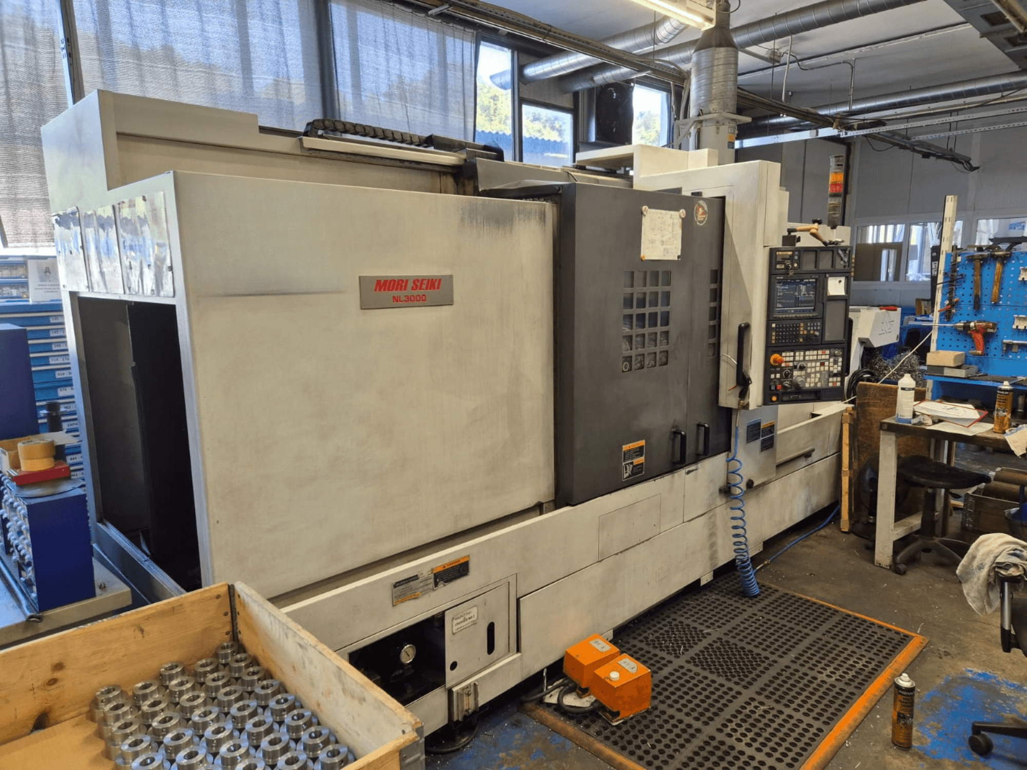 Токарний верстат з ЧПУ Mori Seiki NL3000 в майстерні, вид збоку, на якому видно панель управління, інструменти та металеві деталі в дерев'яному ящику.