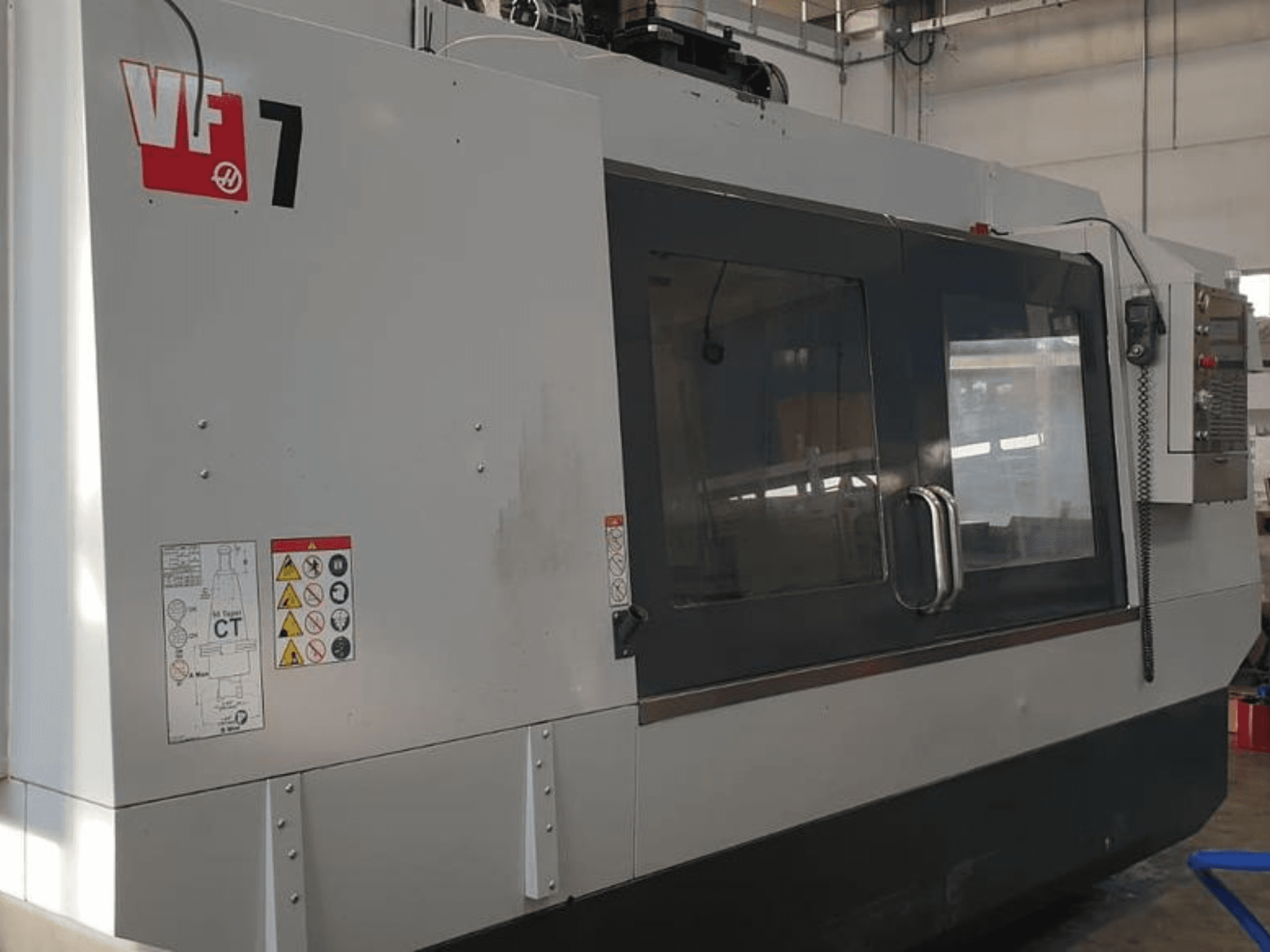 Верстат з ЧПК Haas VF-7 у вигляді збоку, демонструючи металеву сіру обробку, панель керування та етикетки безпеки.
