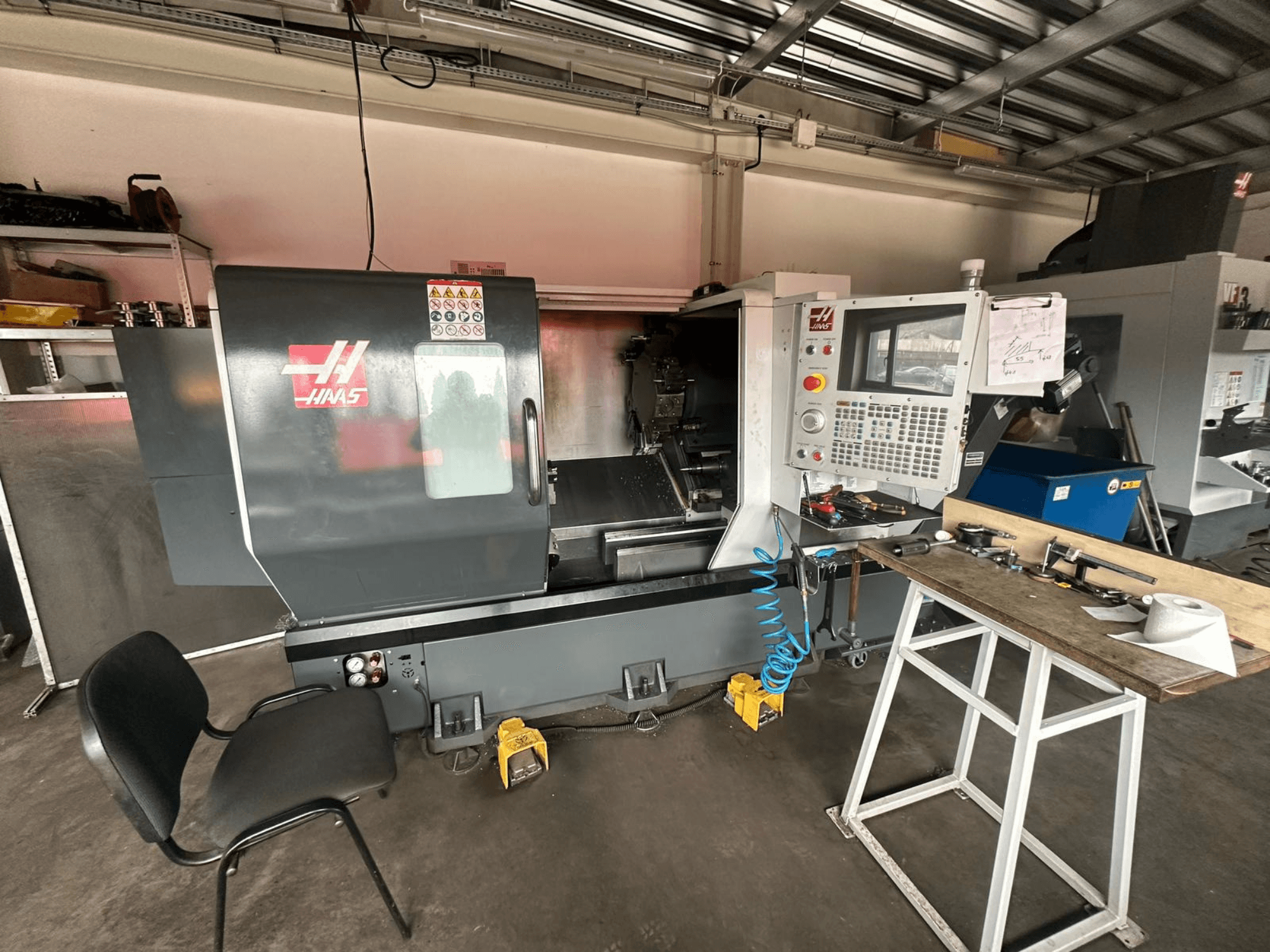 Токарний верстат з ЧПК Haas (модель ST-10) у майстерні, вид збоку: елементи керування, інструменти на робочому столі та стілець поруч.