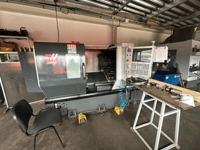 Токарний верстат з ЧПК Haas (модель ST-10) у майстерні, вид збоку: елементи керування, інструменти на робочому столі та стілець поруч.